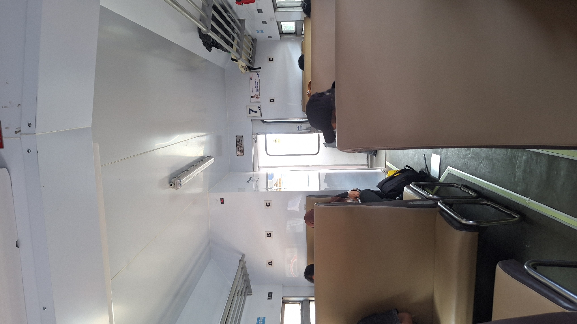 The image shows the interior of a train carriage. Seats are upholstered in beige. Overhead racks are present. Passengers are seated, and one appears to be lying down. Personal belongings, including a black backpack and a laptop, are visible. The letters "A," "B," "C," "D," and "E," are visible on the wall, likely marking seat locations. A door is visible with a sign. Given the specification, the location is Cikarang, Indonesia, though there is no identifying evidence visible in the photo.