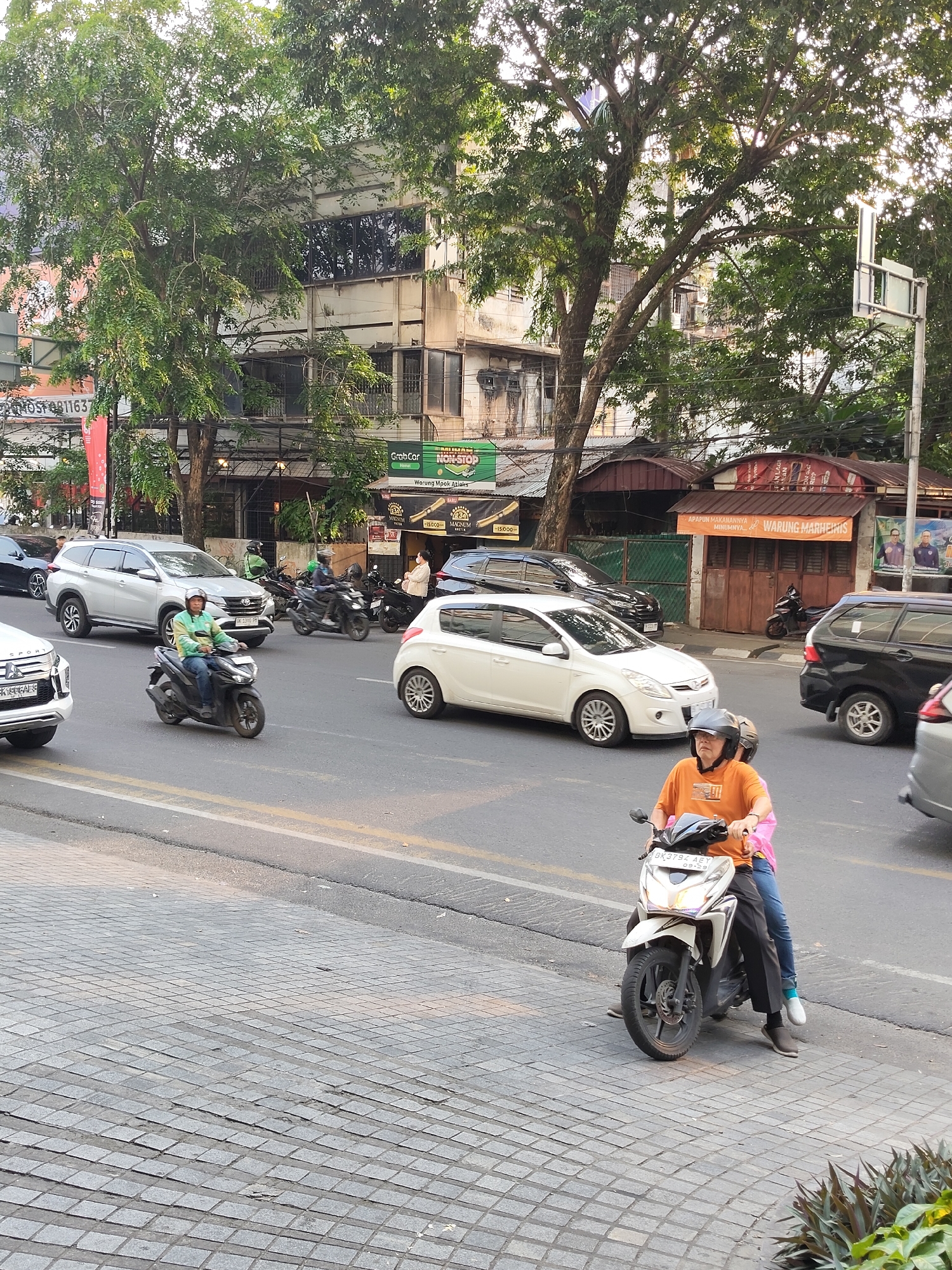 A street scene in Medan, Indonesia, captures traffic and pedestrian activity on a paved road with a sidewalk made of interlocking square pavers. In the foreground, a person in an orange shirt rides a white scooter, accompanied by a passenger wearing a pink shirt. Several other scooters and cars are visible on the road, including a silver SUV, a white sedan, and a black SUV. Buildings with storefronts line the street, with one displaying a sign for "WARUNG MARHEINIS". Trees with dense foliage are interspersed among the buildings, and power lines are visible overhead. The sky is bright, suggesting daylight.