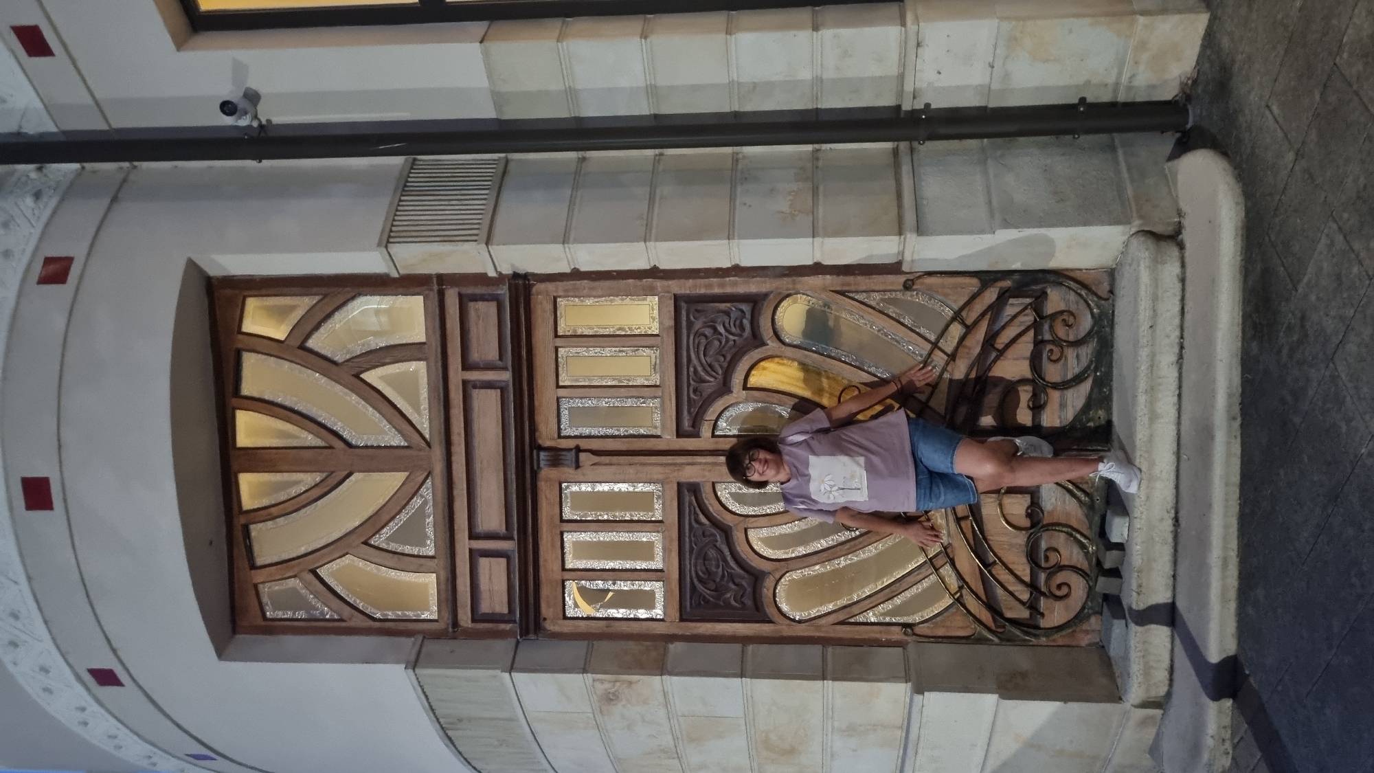 A full-body outdoor medium shot shows a woman standing in front of a large ornate wooden door with glass panels, possibly an entrance to a building in Berehove, Ukraine. The woman, who appears to be of middle age, has short, dark hair and glasses. She is wearing a light purple t-shirt with a white graphic and blue denim shorts. Her white sneakers are visible. She is standing on a concrete step, with her hands resting on the door's decorative metalwork. The door is tall and features intricate carvings and stained-glass elements. The building facade is made of light-colored stone blocks. To the right of the door, a dark pole extends vertically, with a security camera mounted on it. The lighting suggests it is evening.