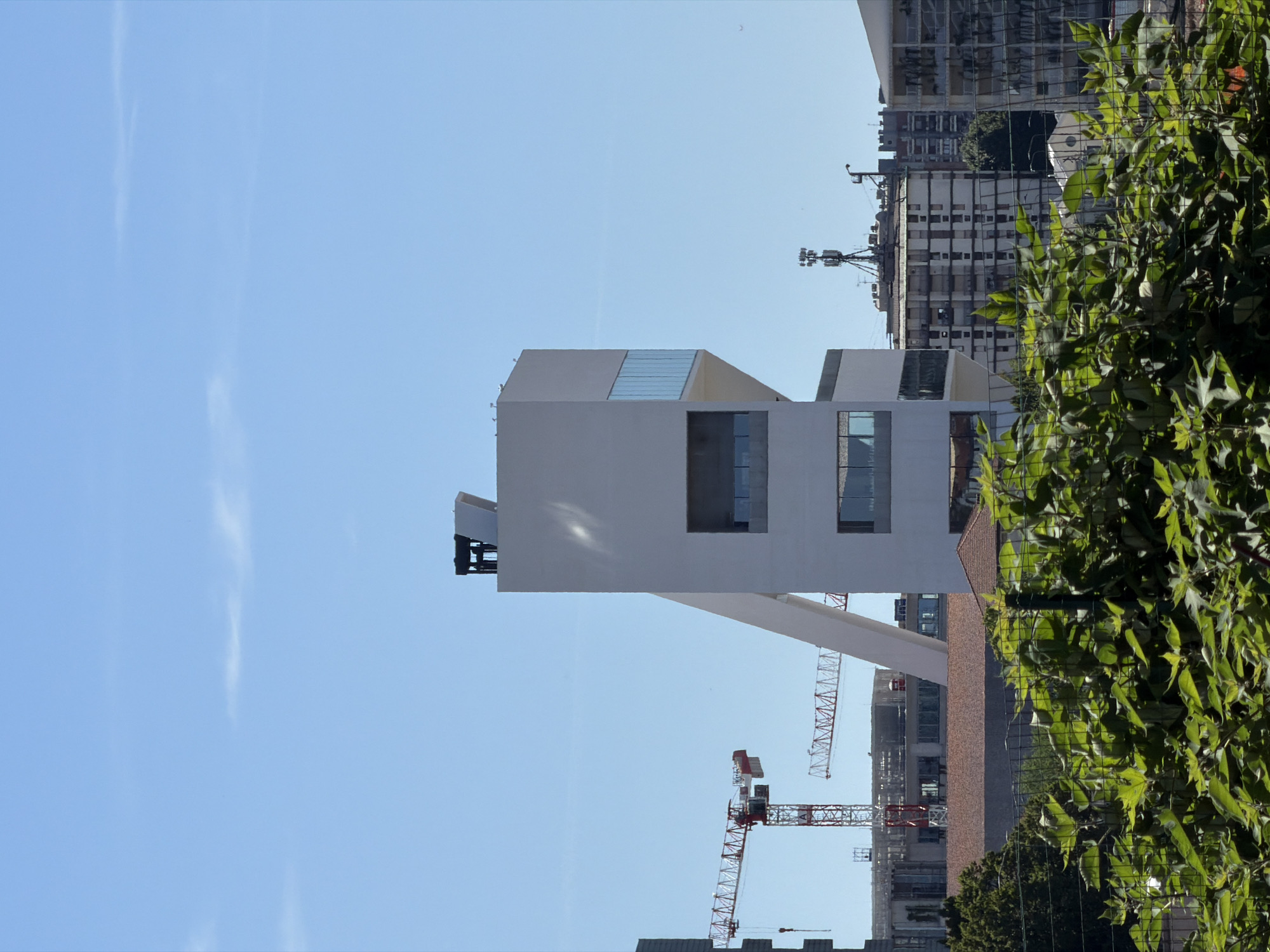 The image shows a view of the Fondazione Prada complex in Milan, Italy, on a clear day. In the foreground, lush green foliage is visible, partially obscuring the view. Behind the foliage, the distinctive "Bar" building of the Fondazione Prada is prominent, a tall, angular white structure with glass windows and an inclined white ramp. In the background, several construction cranes are present, indicating ongoing development in the area. Other buildings, including residential structures and telecommunications towers, are also visible in the urban landscape. The sky is a vibrant blue with a few wispy clouds. There are no people visible in the image.