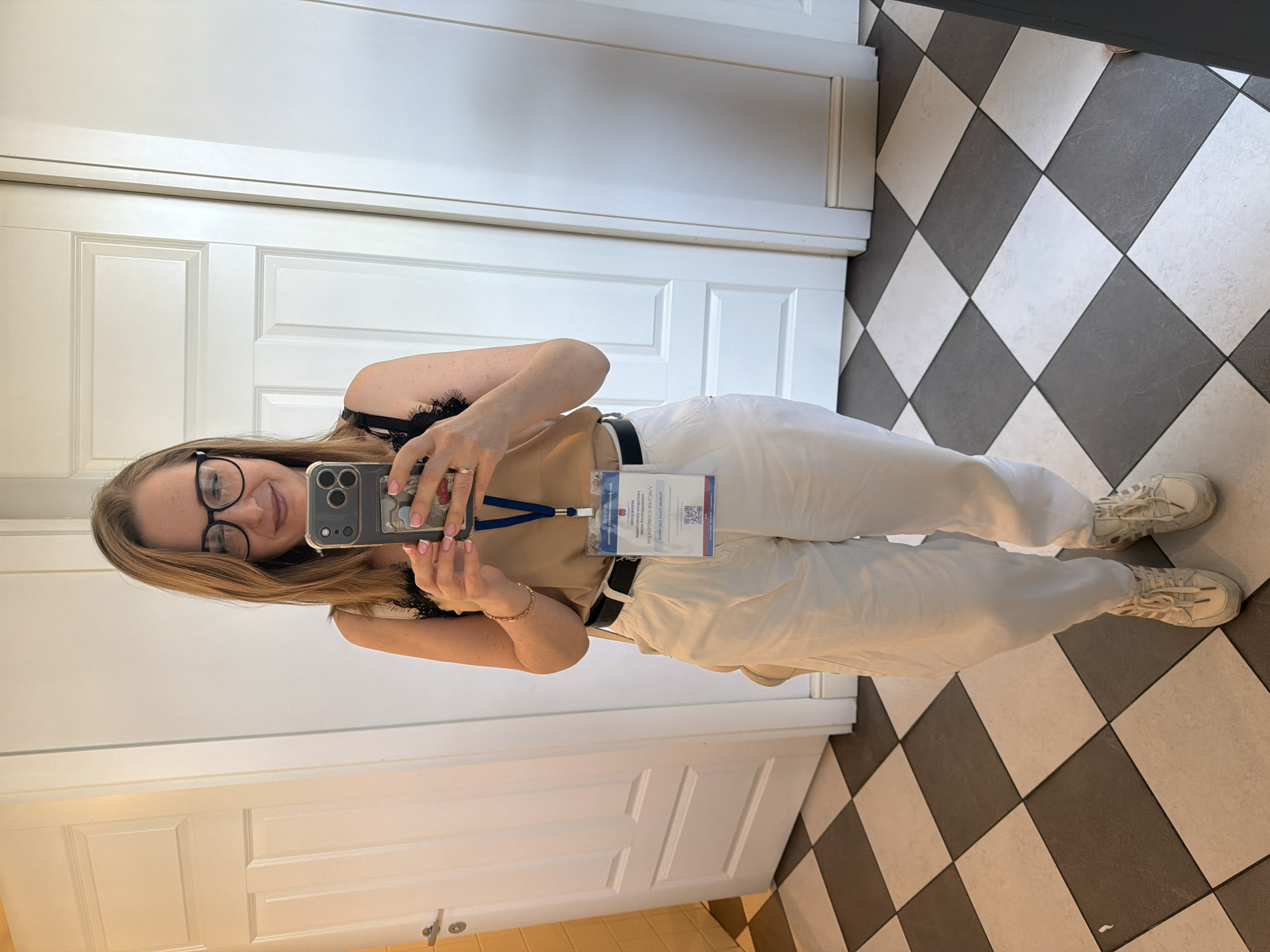 A woman is taking a selfie in a room with white doors and black and white checkered flooring. She is wearing glasses, a beige top, white pants, and white sneakers. A lanyard with a badge hangs around her neck. The location might be a hotel or office space in Moscow, Russia, given the architectural details.