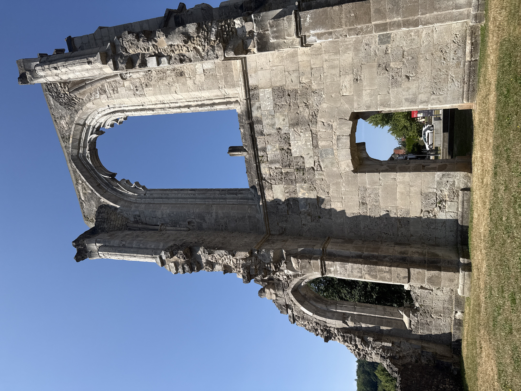 This is a low-angle, full-shot, outdoor photograph of the exterior ruins of the Walkenried Abbey in Walkenried, Germany. The ruins are made of light-colored stone and are crumbling, with large openings in the walls and roof. The sky above is clear and blue. In the foreground, there is a patch of dry grass. Through one of the archways in the ruins, a white car and some buildings can be seen in the background. The image captures the historical and weathered nature of the abbey ruins.