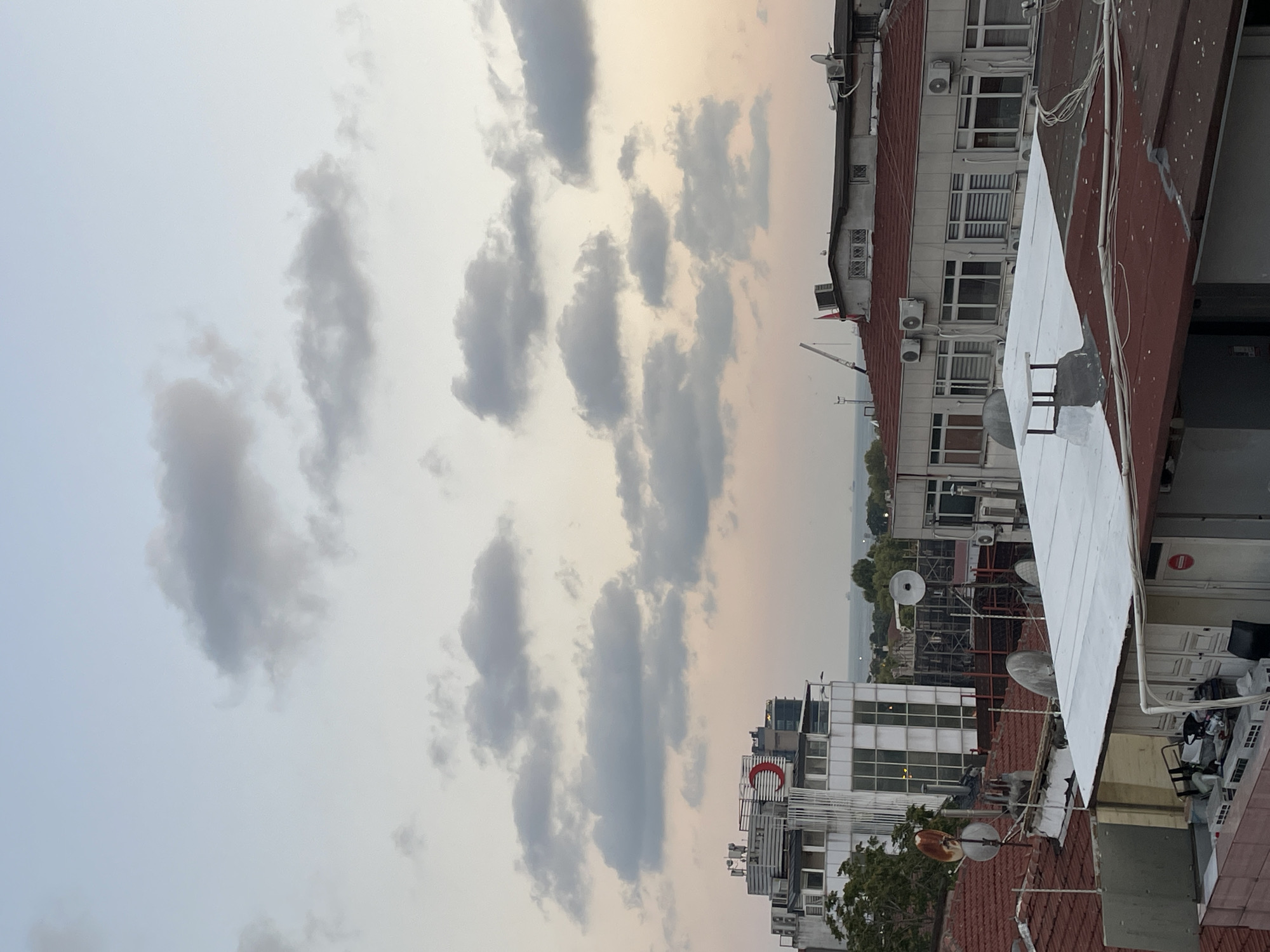 This image captures a view of the urban landscape of Kadıköy, Turkey, likely from a rooftop. The sky is filled with scattered, fluffy grey clouds against a pale blue and pink gradient, suggesting either dawn or dusk. In the background, a body of water, possibly the Bosphorus Strait, is visible with a few ships on the horizon. The foreground is dominated by the rooftops of several buildings, characterized by red tiles and some with white or grey flat roofs.
Prominently featured is a modern white building with large windows and air conditioning units on its upper floors. A large, distinctive red crescent logo is affixed to the side of this building, indicating a connection to the Turkish Red Crescent or a similar organization. Adjacent to this building, other structures with red-tiled roofs are visible, equipped with numerous satellite dishes and air conditioning units, common features in dense urban environments. Some of these buildings appear to be older, with visible signs of wear and tear on the rooftops. The scene is devoid of any visible people. The overall impression is a typical urban vista with the natural beauty of the sky providing a backdrop.