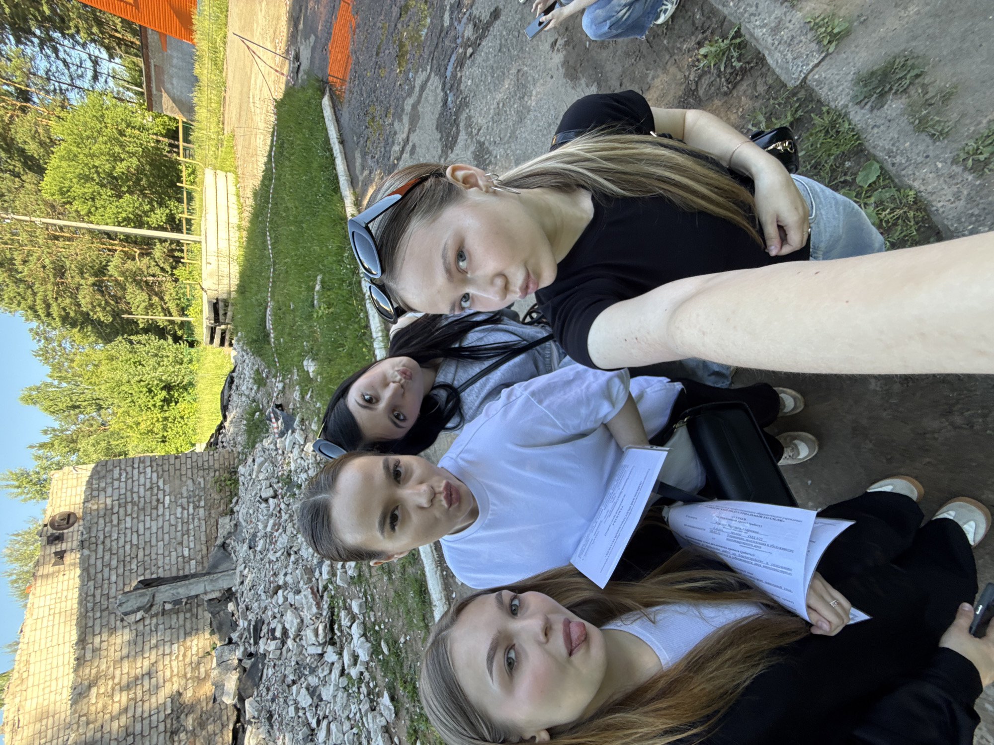 Four young women are taking a selfie outdoors near a partially demolished brick building. The building appears to be constructed of yellow brick and has a large opening with rubble at its base. Behind the building, dense green trees form a backdrop under a clear sky. The ground in the foreground is a mixture of asphalt and dirt, with patches of grass. Three of the women are in the foreground, and the fourth is partially visible in the background. The women are dressed casually in t-shirts and jeans or pants, with sunglasses on two of them. One woman in the foreground is sticking her tongue out, while the other two are puckering their lips. They are holding papers, possibly for a school project or event. The presence of construction debris and the state of the building suggest an industrial or abandoned area. This location is likely in the city of Chaikovsky, Russia, given the context provided.