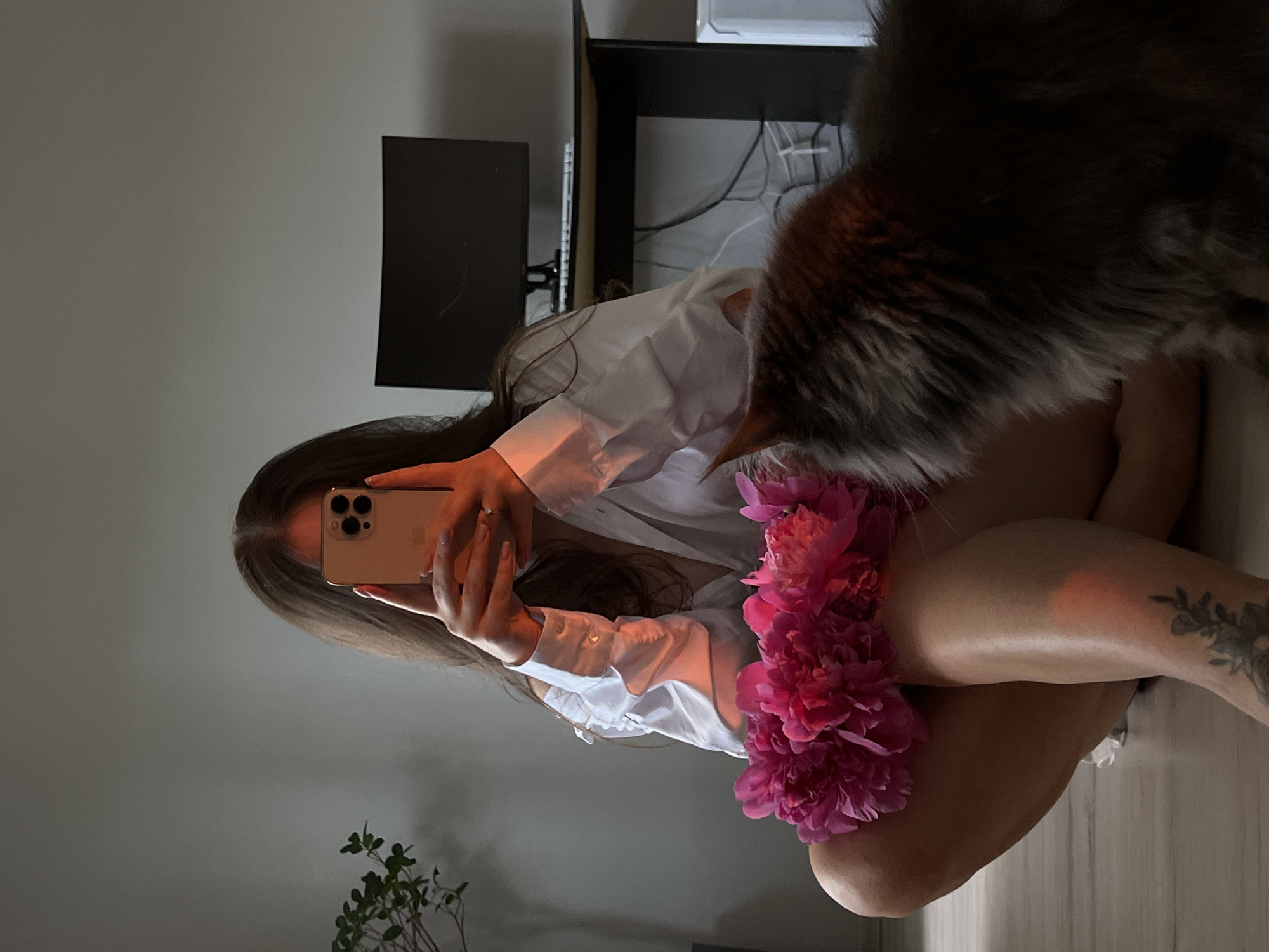 The image shows a woman taking a mirror selfie with her phone, holding a bouquet of pink peonies. She is wearing a white unbuttoned shirt. A fluffy gray cat is in the foreground, looking towards the woman. In the background, a computer monitor is visible on a desk, and a plant is on a shelf. The scene appears to be indoors, likely in a residential setting. There is no information in the image to suggest a specific location in Ternopil, Ukraine.