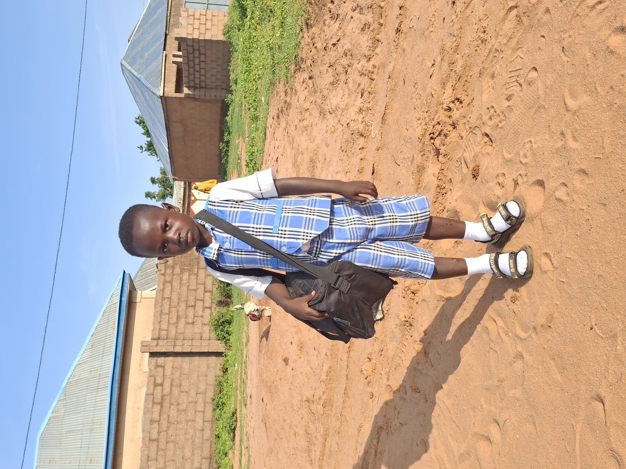 A young boy stands facing forward on a sandy ground in Tumfure, Nigeria, wearing a blue and white plaid uniform with a matching vest and shorts, a white collared shirt, white socks, and dark sandals. He carries a black bag over his left shoulder. In the background, a brick wall separates the sandy area from a grassy patch and two buildings with corrugated metal roofs. A chicken stands to the left of the boy, and another person in a yellow garment is visible in the distance. The sky is clear and blue.
