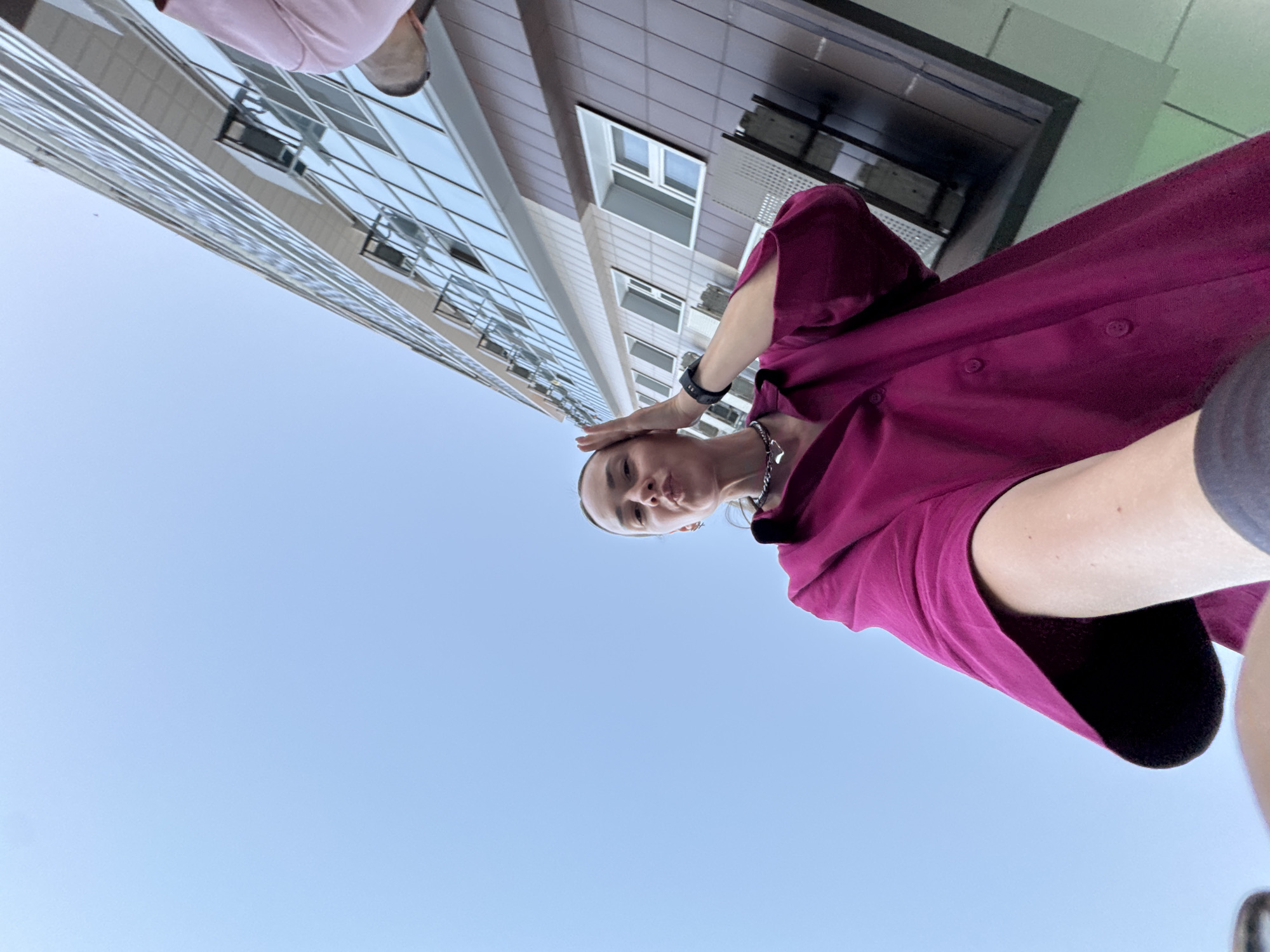 The image is a low-angle, wide-shot selfie taken outdoors. A woman is in the foreground, holding her right hand to her forehead, with her lips pursed. She is wearing a magenta collared shirt and a silver chain necklace. To her right and in the background is a modern building with many glass windows, reflecting the clear blue sky. The scene appears to be in an urban environment, possibly the city of Sochi, Russia, given the architectural style of the building and the clear sky.
