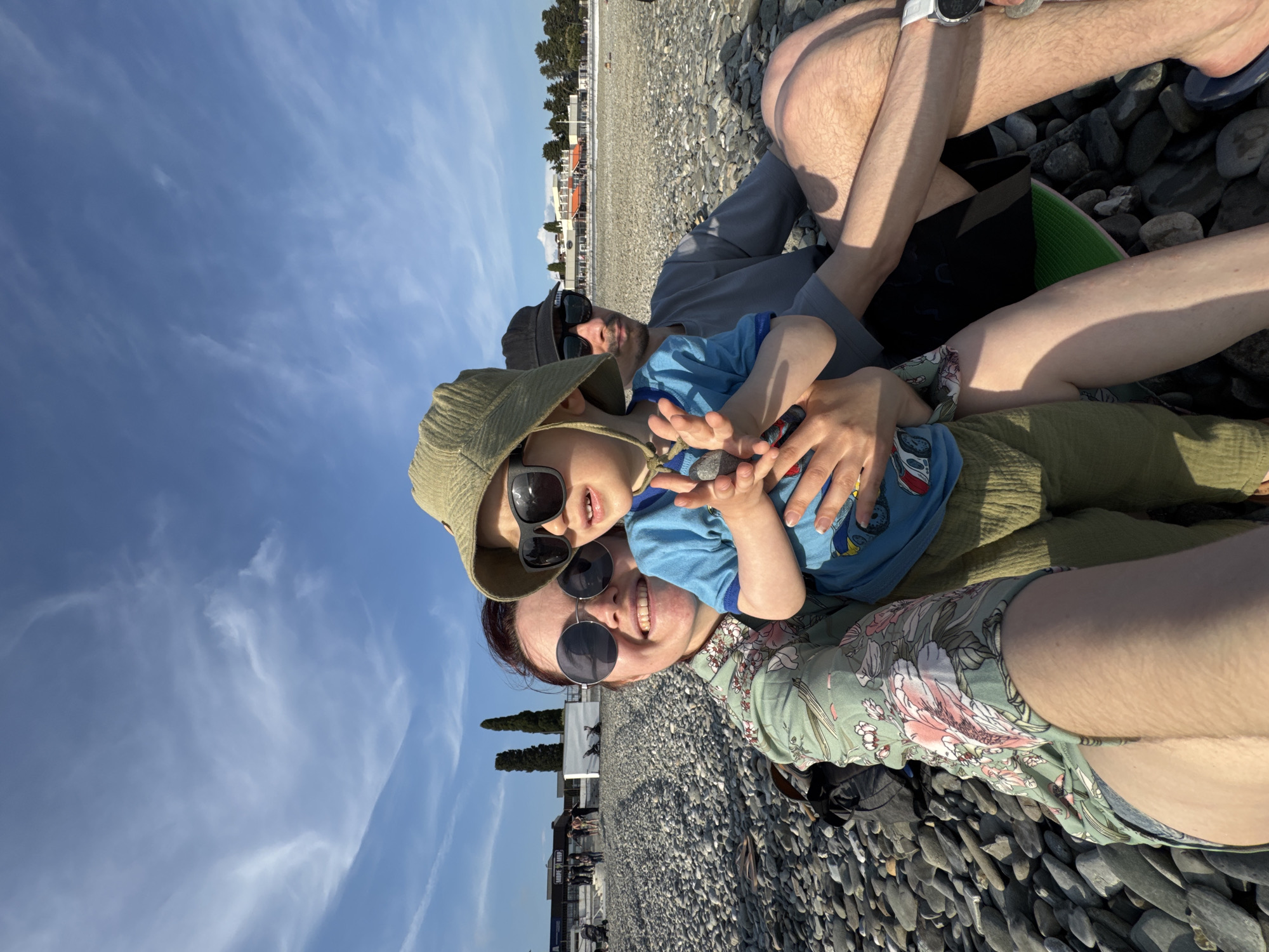 A sunny day at a pebbled beach in Sochi, Russia, features a family lying on the ground. A woman with dark hair and sunglasses is in the foreground, wearing a floral-print shirt and shorts. A young child with sunglasses and a hat sits in her lap, extending his hands. Behind them is a man also wearing sunglasses and a hat. A person's legs and a smartwatch are in the top right corner. The pebbled beach extends into the distance, where buildings and trees are visible. A blue sky with light clouds is overhead.