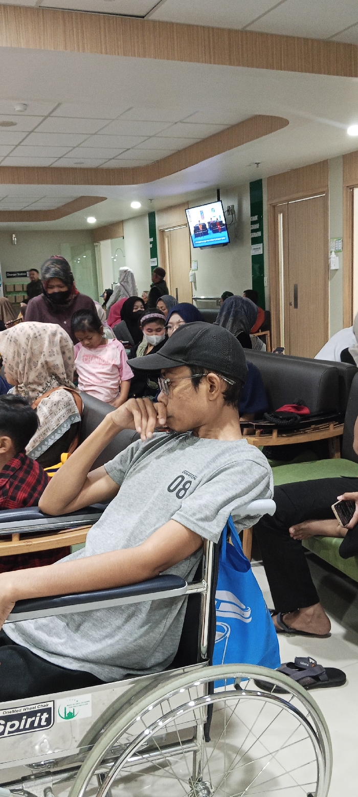 The image shows a man in a gray shirt sitting in a wheelchair in the waiting area of a hospital in Soreang, Indonesia. The man is wearing a black cap and glasses. The man is looking down, and his left hand is resting on his chin.  He has his right arm resting on the armrest of the wheelchair.  There are several other people in the background of the photo. They are mostly seated on couches and benches. The waiting area is clean and well lit. The walls are painted in a light color. The image is taken from a low angle, making the man in the wheelchair appear large in the image.  The background is blurred. The image is taken in a room with a tiled floor. There is a TV on the wall, as well as a wooden countertop, and other furniture. The image captures a common scene in a hospital waiting area. The man in the wheelchair appears to be waiting patiently, while the other people in the room are also waiting for their turn. The scene is quiet and peaceful. The image is taken in a location with a latitude of -7.032 and a longitude of 107.527.