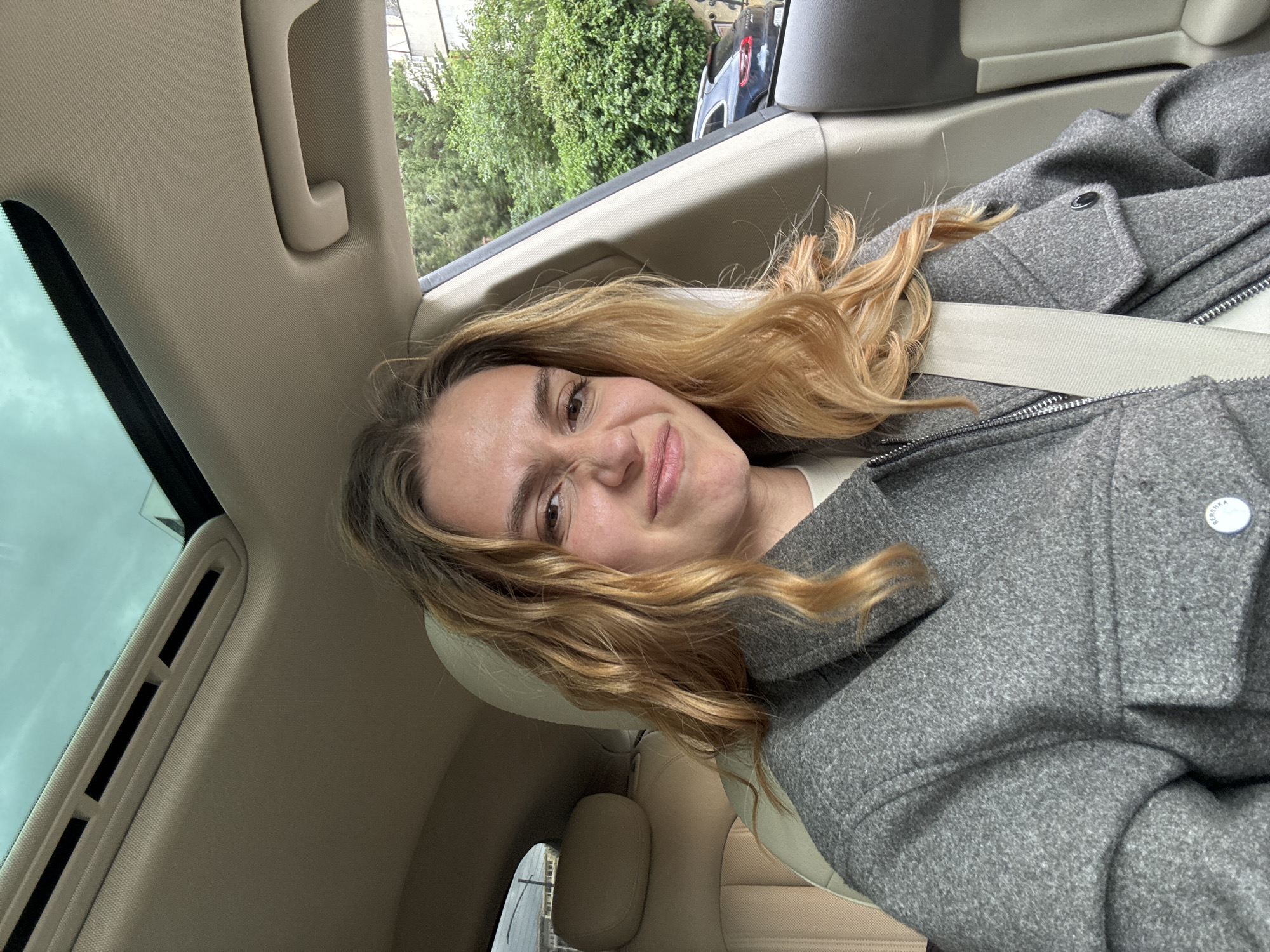 A woman with long, wavy blonde hair is wearing a gray wool coat and a seatbelt in what appears to be the back seat of a car. The interior of the car is beige and includes a headrest. Outside the window, there are green trees. The location is unknown.