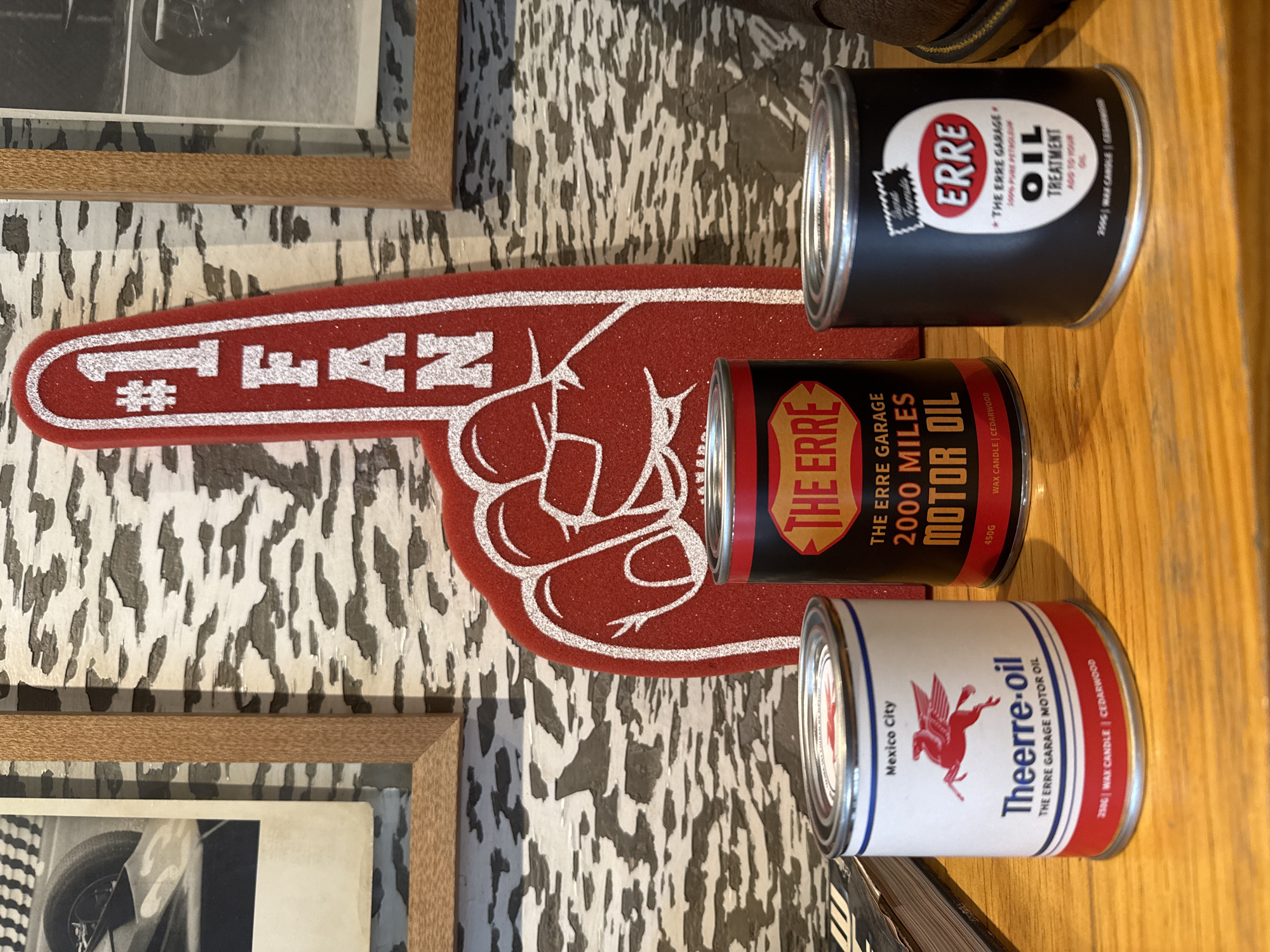 The image shows a close-up of three cylindrical cans stacked on a wooden surface. The cans appear to be oil cans that have been repurposed as wax candles, as indicated by labels containing information about "wax candle" and weights in grams. The bottom can is white with red and blue stripes, and a red illustration of a winged horse. It features the brand "Theerre-oil" and "The Erre Garage Motor Oil", along with the text "Mexico City", indicating a location within the city of Mexico City, Mexico. The middle can is black with red accents. It says "THE ERRE 2000 MILES MOTOR OIL". The top can is black and has the brand ERRE "THE ERRE GARAGE OIL TREATMENT". Behind the cans, there's a red foam hand and a mottled background that appears to be a wall. A framed image can be partially seen to the left of the composition.