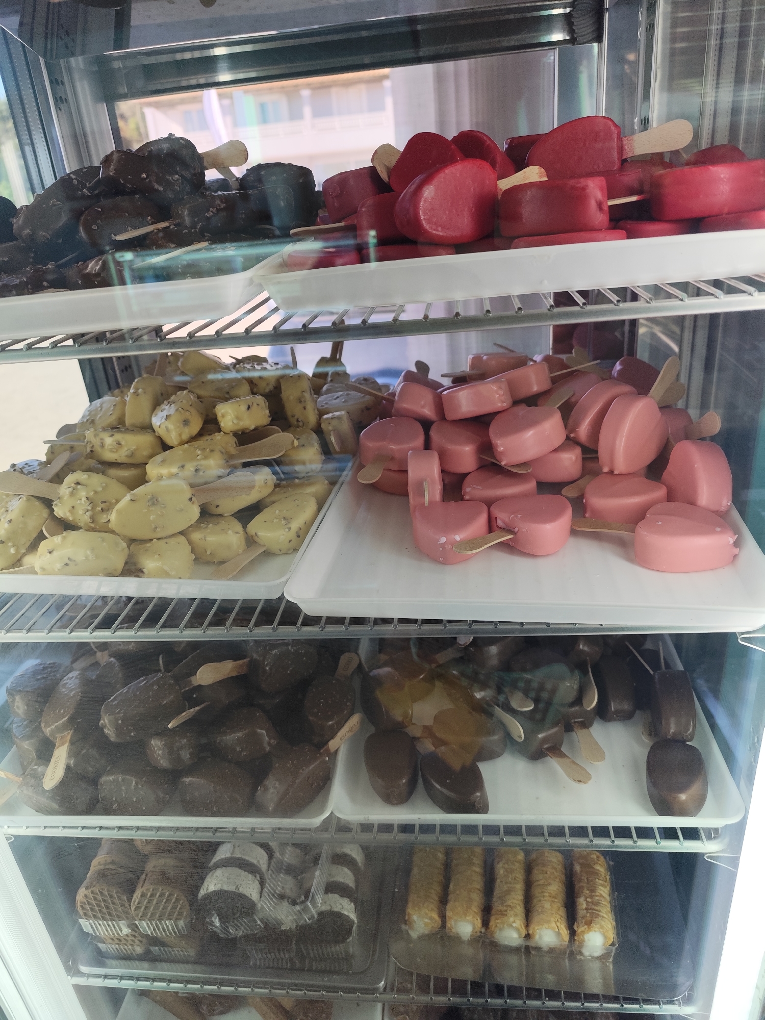 The image shows a refrigerated display case in a store located in the city of Dafni, Greece. The case contains multiple shelves of various ice cream and dessert items on wooden sticks. On the top shelf are chocolate covered ice cream bars on the left and red ice cream bars on the right. The next shelf down has light yellow ice cream bars on the left, and pink ice cream bars on the right. The third shelf has dark chocolate ice cream bars on the left, and chocolate covered ice cream bars on the right. The bottom shelf has waffle cookies on the left and cannolis on the right.