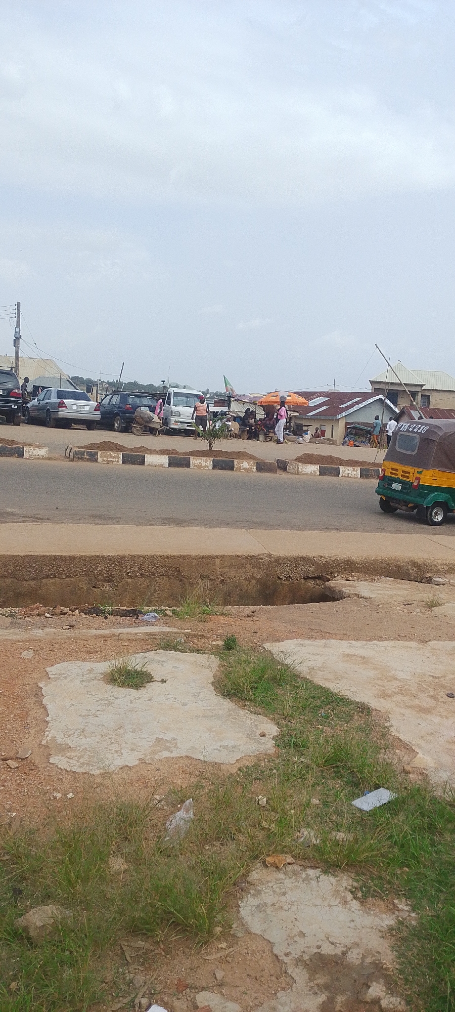 This media file captures a bustling street scene in Gura Top, Nigeria, under an overcast sky, suggesting it might be morning or late afternoon. The foreground shows a weathered concrete slab with sparse grass growing through it, adjacent to a raised curb and a road. The middle ground reveals a lively market area with people gathered, some tending to stalls under colorful umbrellas. Several vehicles are parked or driving on the road, including cars, a white van, and a distinctive yellow and green auto-rickshaw (Keke Napep) with the license plate "YTA-224D" visible.
In the background, more buildings with corrugated iron roofs and some shops line the street. The activity appears to be a typical day of commerce, with individuals interacting, selling goods, and moving about. The overall impression is one of a busy, local marketplace integrated with road traffic. There are no strong emotional cues visible, but the density of people and vehicles suggests a dynamic and active environment. The presence of the auto-rickshaw and the architecture are strong indicators of a Nigerian location.