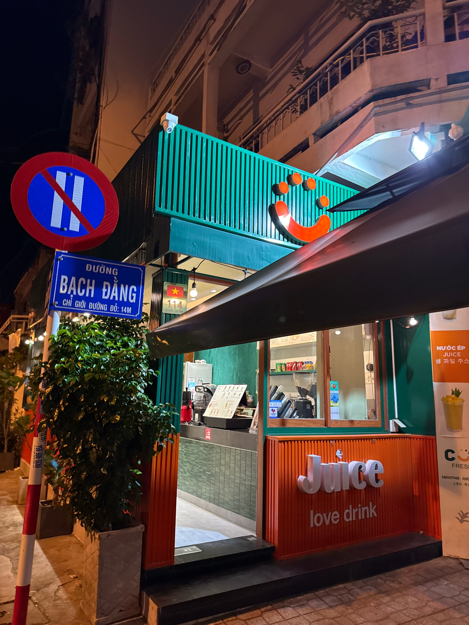 An exterior shot at night depicts a juice bar called "Juice love drink" in Nha Trang, Vietnam. The bar has a vibrant orange facade with the name "Juice" in large, white, raised letters and "love drink" below it. A smiling smiley face with three dots is visible above the word "Juice". A green, slatted awning extends from the building. To the left of the bar, a street sign reads "ĐƯỜNG BẠCH ĐẰNG" (Bach Dang Street) with smaller text indicating a road limit. A dense green bush is planted in a grey, textured pot next to the sign. A traffic sign with a red circle, blue center, and a red diagonal line crossing two vertical white lines is also present, indicating "no parking." A security camera is mounted on the green structure. Inside the bar, visible through a window, are shelves stocked with drinks and what appears to be a coffee grinder. A Vietnamese flag is displayed on a small sign next to the entrance. A promotional poster for "Nước ép" (juice) is on the right side of the building. The ground in front of the bar is paved.