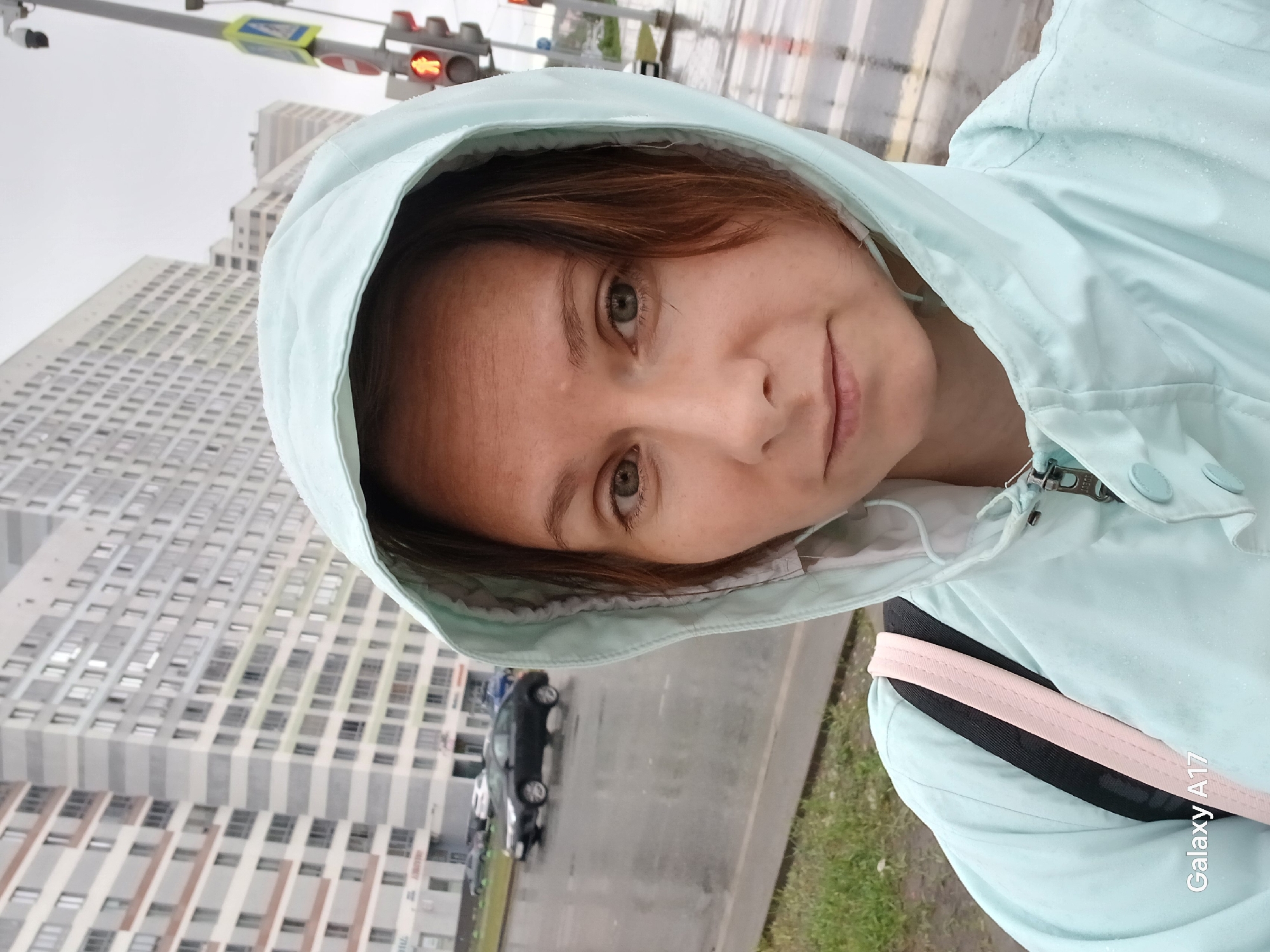 A woman is taking a selfie in what appears to be a rainy urban environment. She is wearing a light blue hooded jacket. Buildings are visible in the background. A street with cars and traffic lights is also discernible. The buildings in the background are typical of modern city architecture. There is a "Galaxy A17" watermark in the lower right. Considering the details of the buildings, street design, and overall layout, the location is potentially Yekaterinburg, Russia.