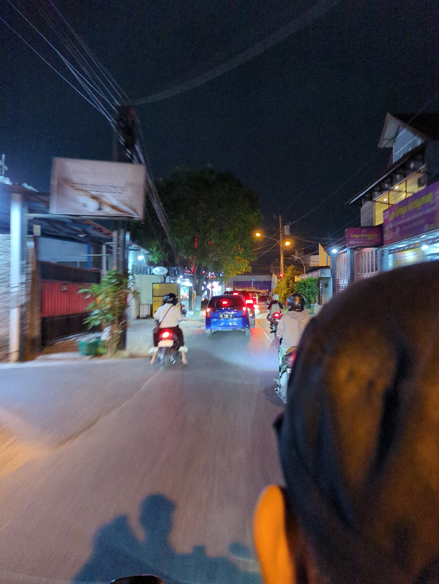 The image depicts a street scene at night in Gentan, Indonesia, likely on Jalan Raya Gentan. In the foreground, the back of a person's head and shoulder, wearing a dark helmet and possibly a light-colored jacket, is visible, suggesting the photo is taken from the perspective of someone riding on a motorcycle. Several motorcycles and a blue car are moving along the asphalt road, illuminated by streetlights and the vehicles' headlights. On the left side of the road, there are buildings with signs and what appears to be a billboard. Trees line the right side of the street, interspersed with more buildings. The sky is dark, indicating nighttime. The overall impression is that of traffic on a residential or commercial street during the evening.