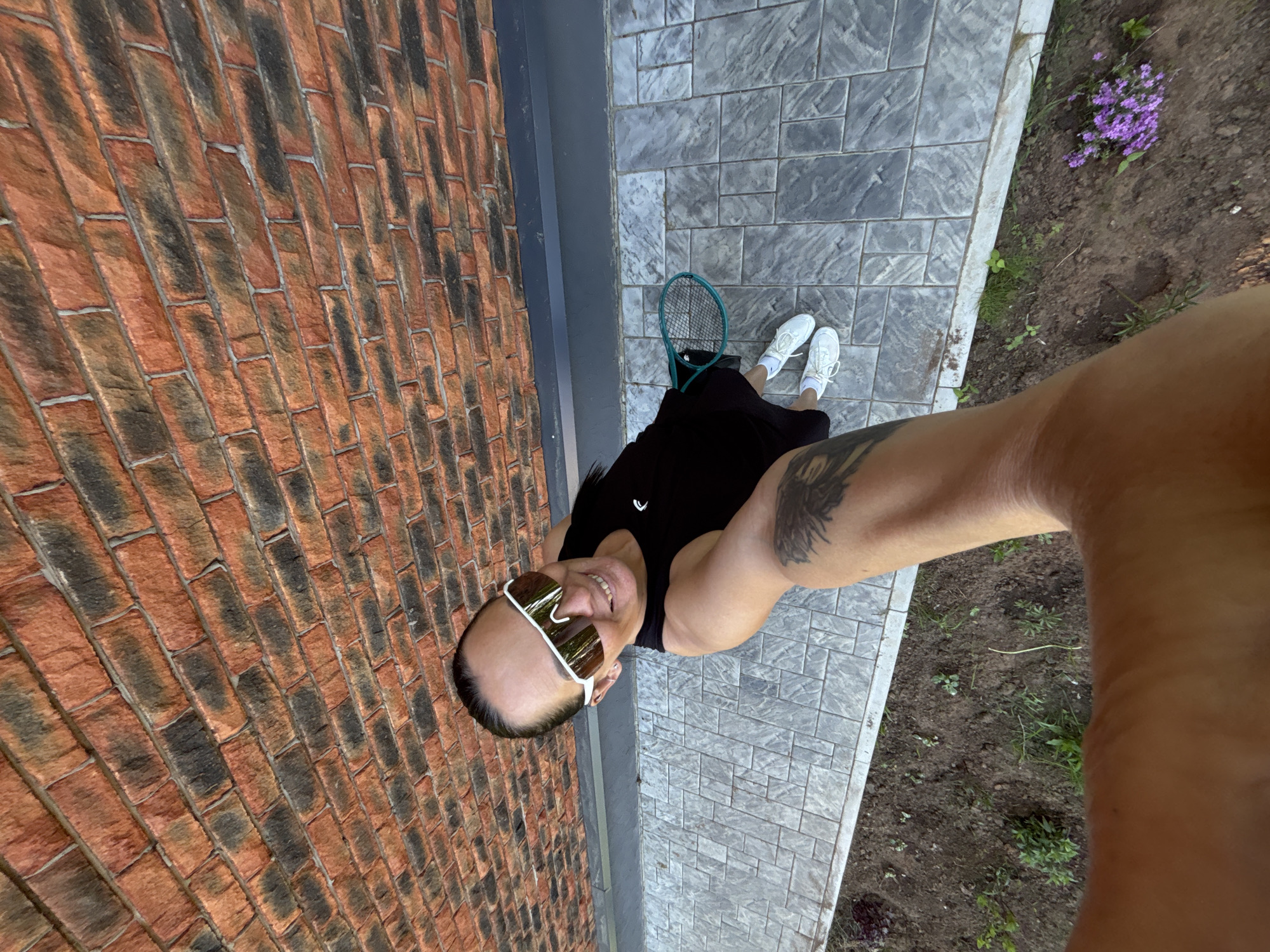 A selfie, taken from a high angle, shows a person standing on a paved area near a brick wall in Chaikovsky, Russia. The person, wearing sunglasses, a black top, and white shoes, is holding a tennis racket. Some small purple flowers grow near the edge of the paved area, next to a dirt area. A tattoo is visible on the person's arm.