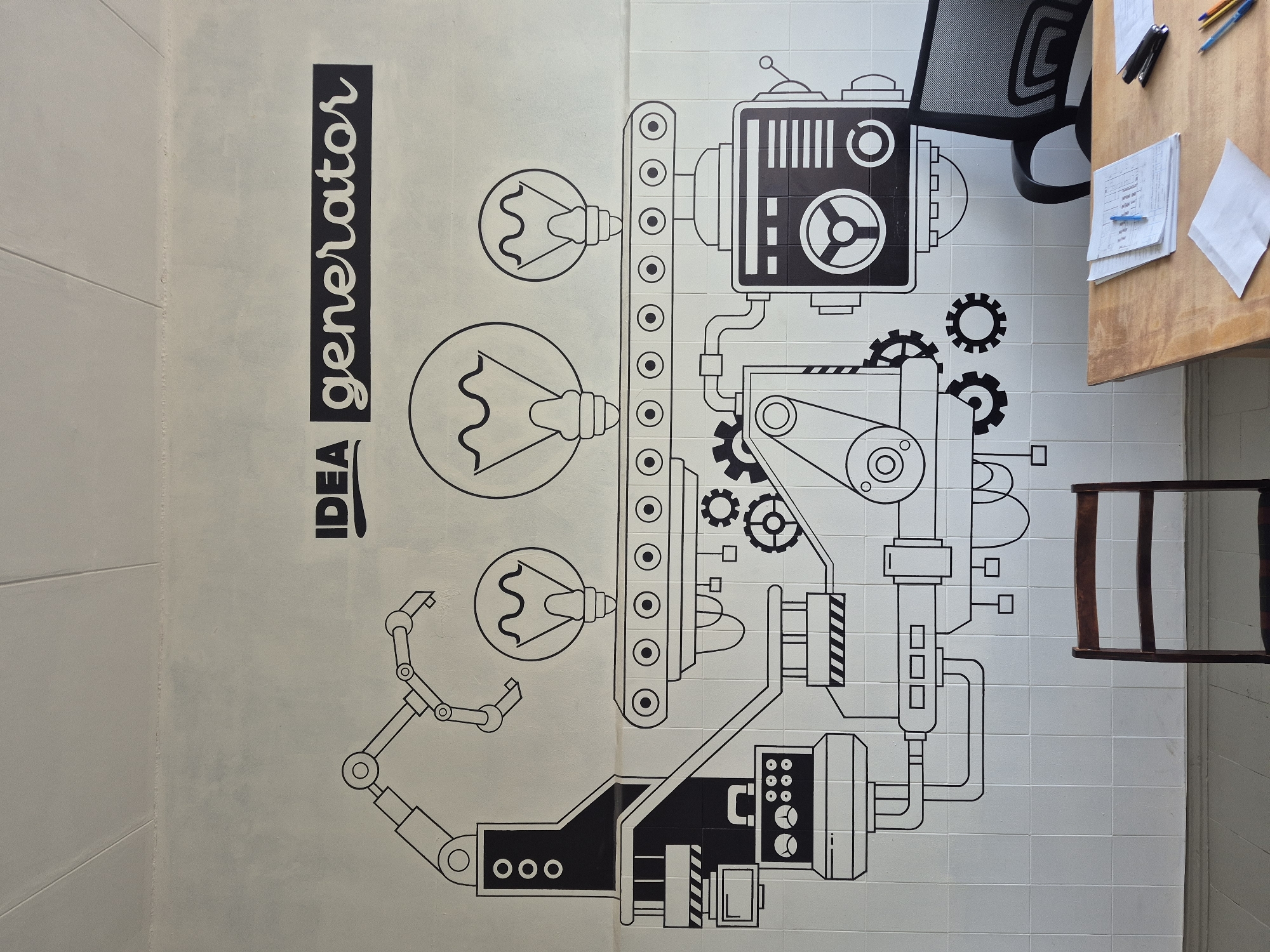The image shows a wall with a drawing of an "Idea Generator" machine on it. The machine consists of various mechanical parts, including light bulbs, a conveyor belt, gears, and robotic arms. The drawing is in black and white and appears to be made on tiles. Adjacent to the wall is a wooden table with papers, a stapler, and pens on it. There are two chairs, one with a mesh back and the other made of wood. The location is unknown.
