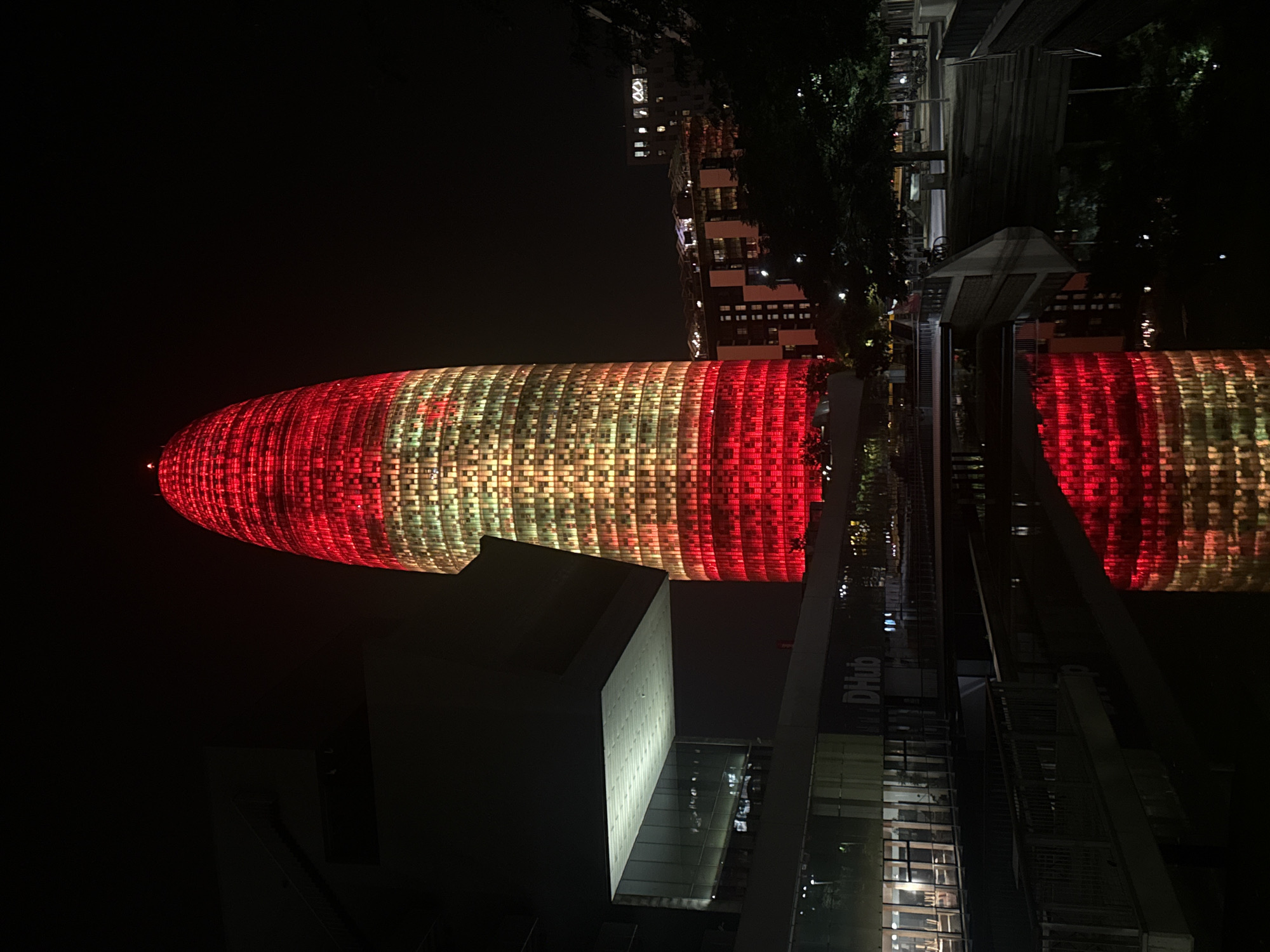 The image shows the Torre Glòries skyscraper in Barcelona, Spain at night, illuminated in red and yellow lights. The skyscraper is a prominent landmark in the city's skyline, located in the Poblenou district. In the foreground, a dark body of water reflects the illuminated building, creating a symmetrical effect. Adjacent to the skyscraper, a modern building with a distinctive cantilevered section is visible. The surrounding area appears to be a public space with walkways, trees, and other buildings. The overall scene depicts a nighttime cityscape, highlighting the architectural beauty of the Torre Glòries and its surroundings.