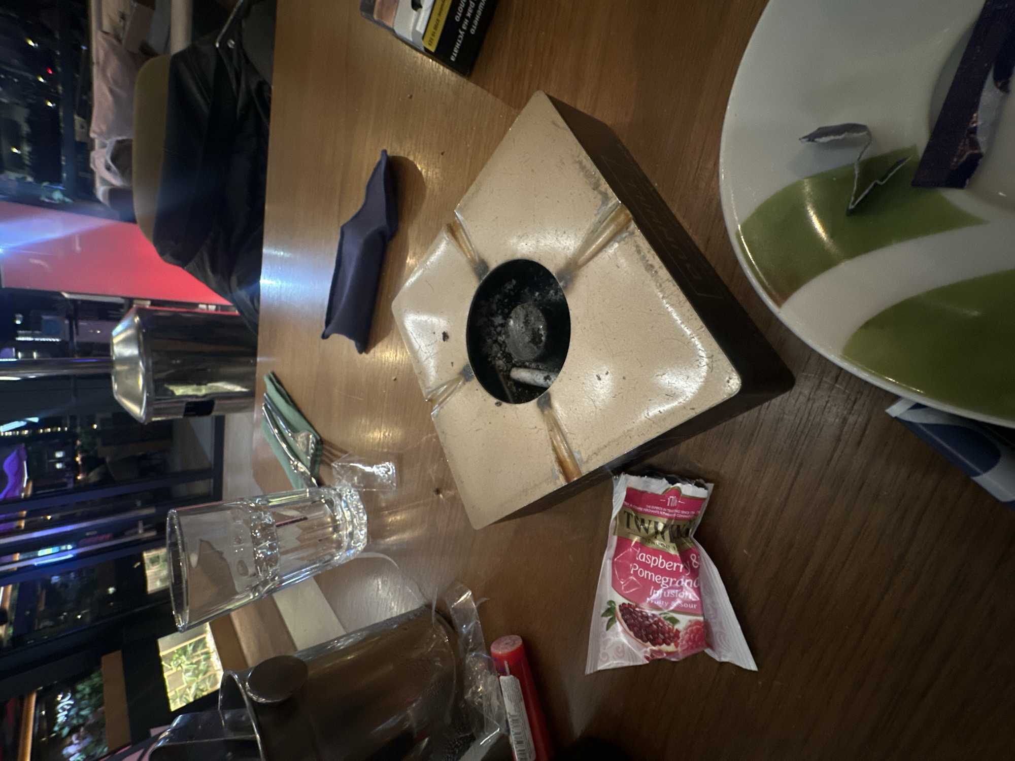 A table in a café in Sofia, Bulgaria, displays various items including a square ashtray filled with cigarette remnants, a packet of Twinings Raspberry & Pomegranate Infusion tea, a glass of water, cutlery, a plate with a packet on it, and personal items. The scene suggests a person or group was dining or taking a break. Outside, through a window, city lights are visible.