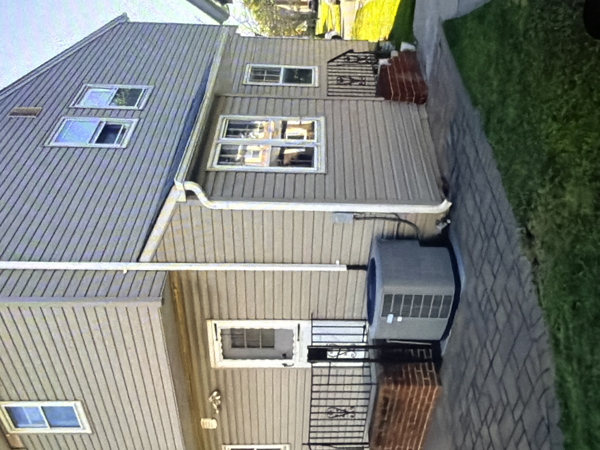 The image shows the exterior of a two-story house, likely in a suburban or residential area. The house has light tan siding and white trim around the windows and doors. There are several windows visible on both floors, some open and some closed. A white door with a screen is on the first floor, with a black railing leading up to it. An air conditioning unit is situated on the patio in front of the house. The patio is paved with interlocking bricks. To the right of the house, there is a small set of brick steps leading to a railing. The area around the house is landscaped with green grass and some trees are visible in the background. The sky is mostly clear with some visible clouds. The image appears to be taken during the daytime, likely in the afternoon given the lighting. There are no people or animals visible in the image.