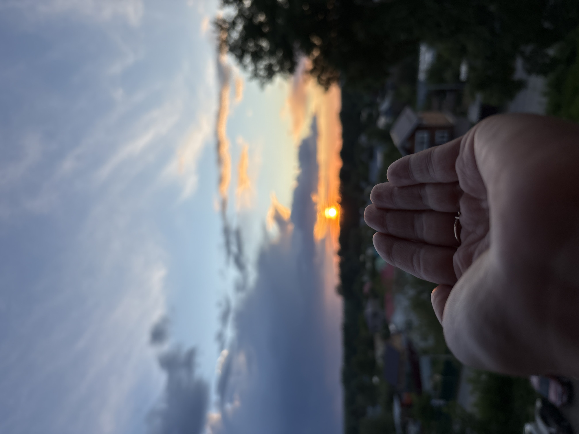 The image captures a sunset partially obscured by a right hand wearing a ring. The background reveals a cityscape, including residential buildings and foliage, indicating a suburban or residential area. The sky is a mix of blue and orange hues with various cloud formations. The picture appears to be taken vertically and then rotated. Based on the visual elements, it seems the location is somewhere near the city of Chaikovsky, Russia, likely from an elevated position offering a view of the surrounding residential landscape during sunset.