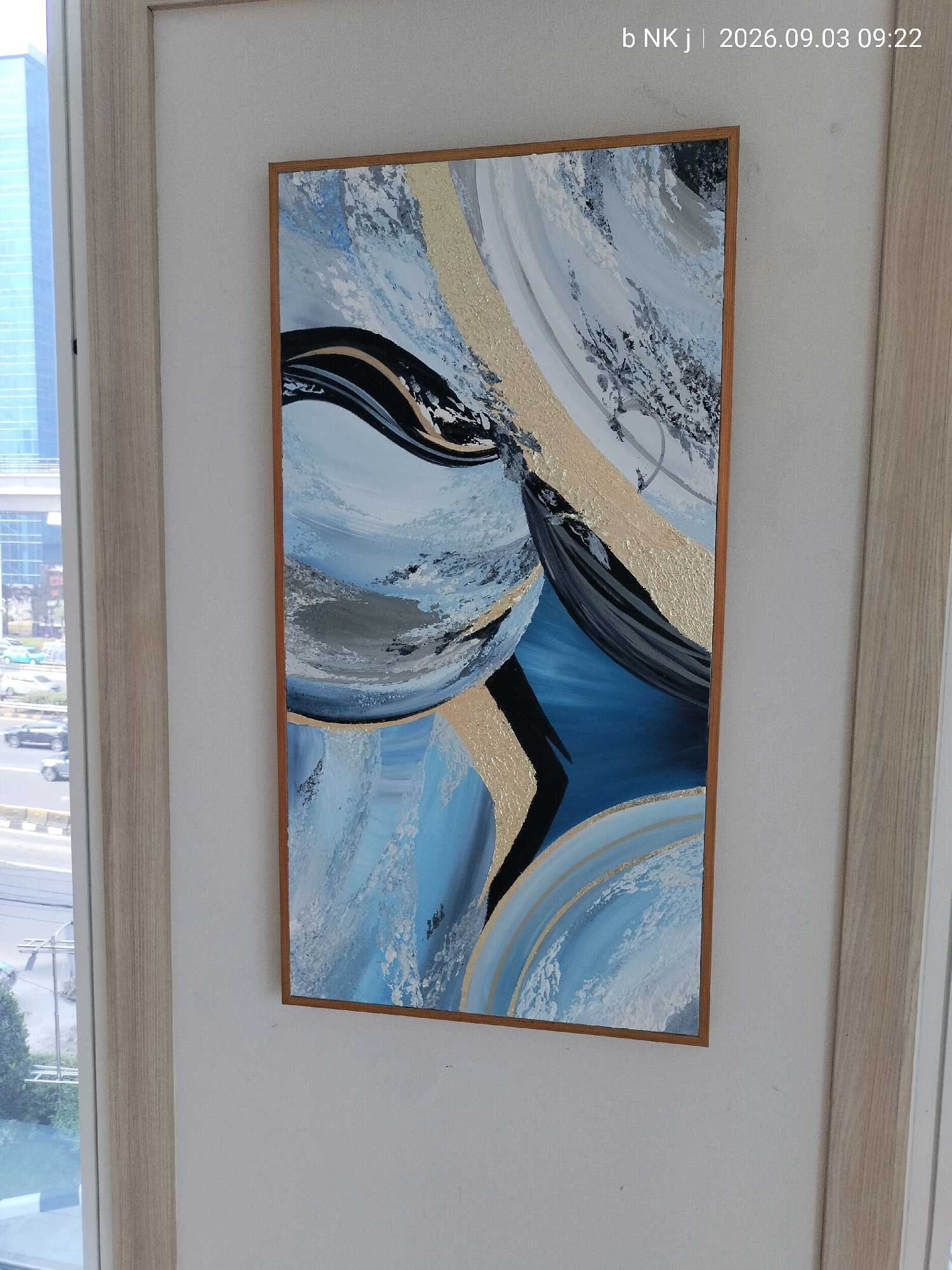 A framed abstract painting is displayed on a wall next to a window with a view of urban scenery. The painting features bold, sweeping strokes of blue, black, white, and metallic gold. The window overlooks a street with cars and buildings, suggesting an urban setting, possibly South Jakarta, Indonesia, given the watermark "b NK j | 2026.09.03 09:22" which appears to be a timestamp or identifier. The wall is painted a light color, and the frame of the painting is a light wood. The overall impression is one of modern interior decor in a city environment.