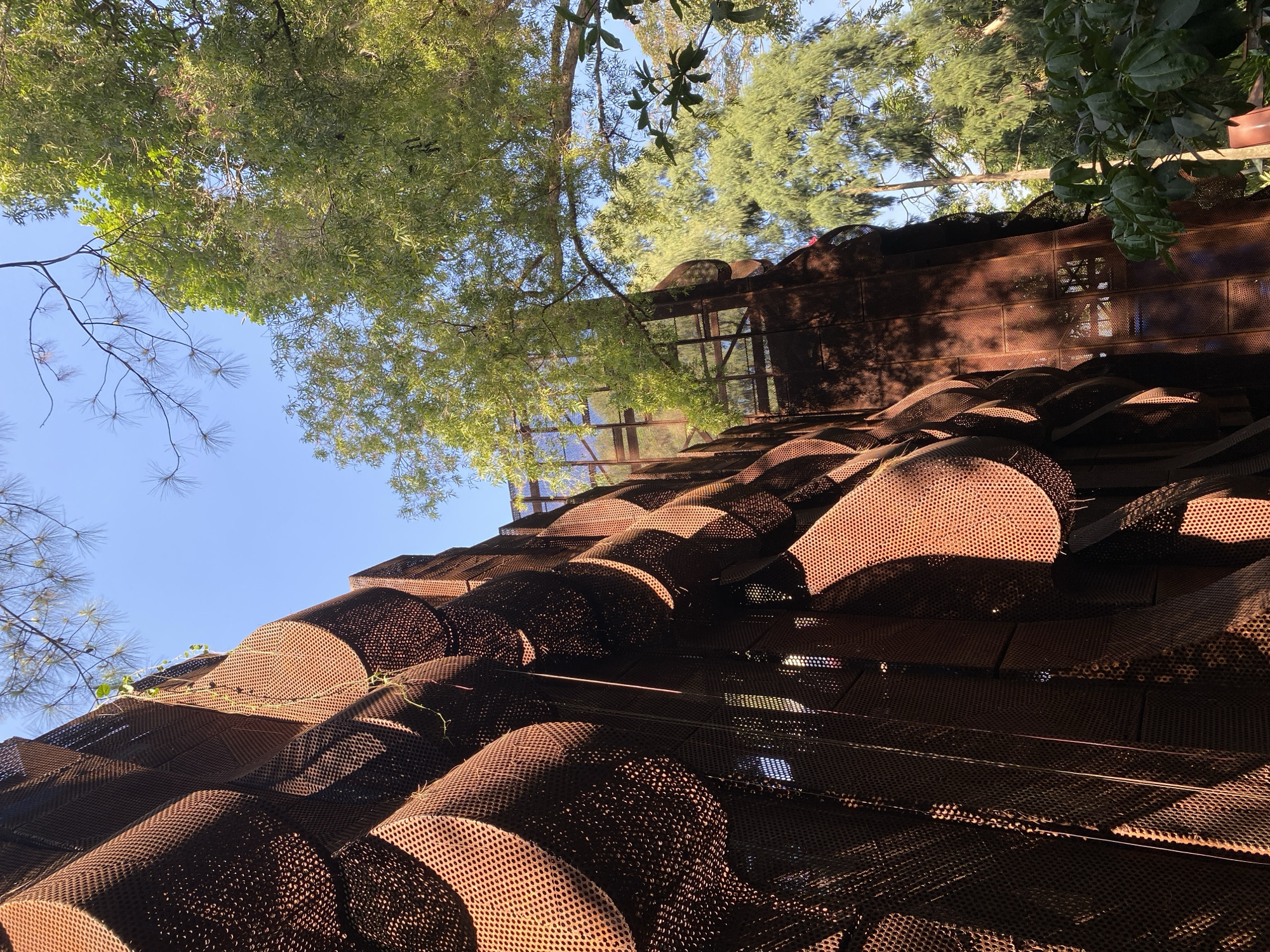 The image captures a unique architectural structure in the city of Ciwaruga, Indonesia. The structure is made of rusted metal panels that form an undulating, textured facade. These panels have a perforated pattern, creating a play of light and shadow. The perspective is from below, looking up towards the sky and through the structure. Green trees and foliage are visible around and above the building, suggesting a natural or park-like setting. The sky is clear and blue, indicating a sunny day. There are no people visible in the image. The overall impression is one of an artistic and organic-looking building integrated with its natural surroundings.