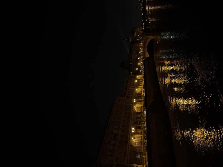 A nighttime scene captures the River Seine in Paris, France, viewed from the Right Bank, looking east-southeast towards the Île de la Cité. The prominent structure on the left bank is a long, multi-story section of the Louvre Palace, characterized by a classical facade with numerous arched windows. This building is brightly illuminated by a series of golden streetlights along the quay below. Indistinct vehicles are visible parked or moving on the street adjacent to the building. The dark water of the Seine occupies the lower half of the frame, reflecting the warm, golden light from the streetlamps and buildings, alongside some red and white light reflections from distant sources. A stone arch bridge, likely the Pont des Arts or Pont Neuf, spans the river in the mid-ground. Beyond the bridge, on the Île de la Cité, architectural features consistent with the Palais de Justice and Conciergerie, including a dome and darker spired structures, are dimly visible against the dark horizon. The sky is dark and clear. No individuals are discernible within the frame.