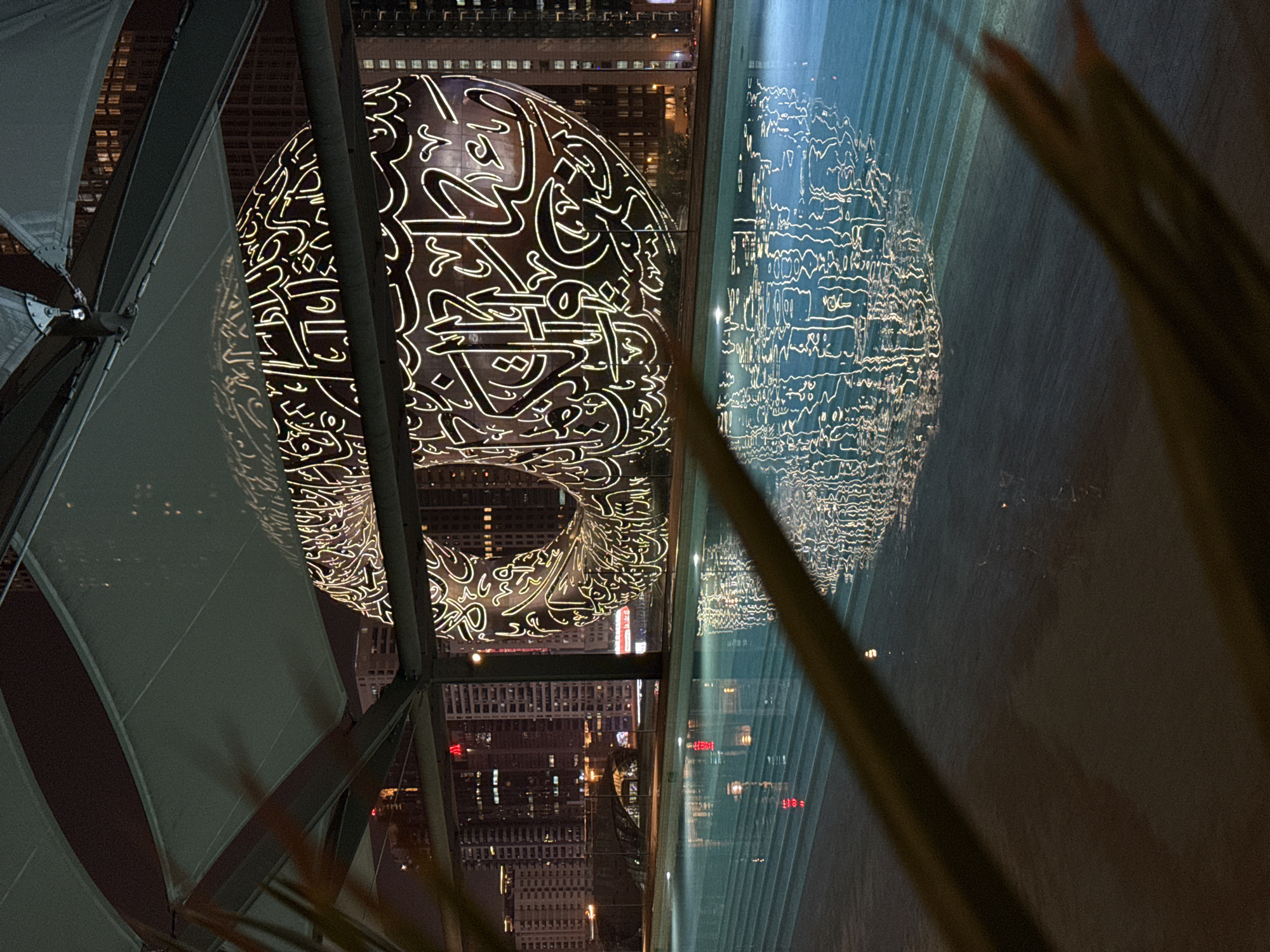 The image depicts the Museum of the Future in Dubai, United Arab Emirates, at night. The distinctive donut-shaped building with Arabic calligraphy is illuminated and reflected in a glass surface. Behind the museum and in the background, there is a cityscape with various buildings and lights. In the foreground, there are architectural details of the structure the image was taken from and vegetation.