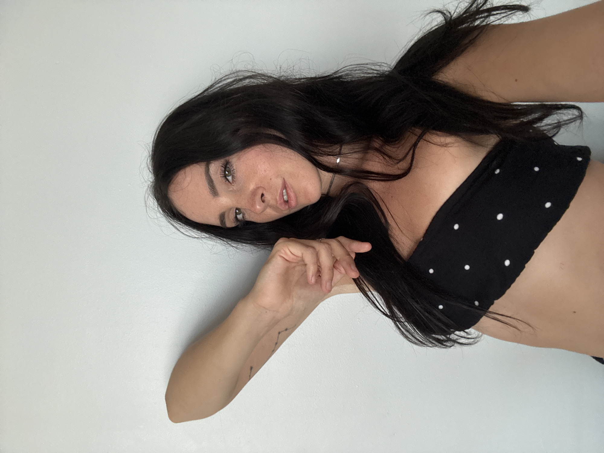 A woman with long, dark hair and fair skin is lying down against a white wall. She is wearing a black strapless top with white polka dots. Her left arm is bent and her hand is resting on her hair, while her right arm is extended behind her. She is looking at the camera with a relaxed expression. The image is a close-up shot, and the background is plain white. There is a faint tattoo on her left arm that appears to be a constellation. The lighting is natural and even, suggesting it is daytime. There are no other objects or people visible in the frame. The setting is indoors, likely a room with white walls.
