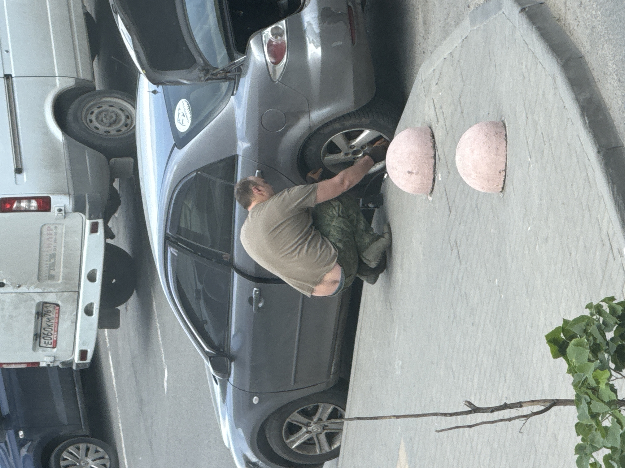 The image depicts a man squatting next to the front wheel of a grey car parked on the side of a street in what appears to be a city. The man is wearing a light brown shirt, camouflage trousers, and appears to be working on the car wheel. The car's trunk is open. Next to the car is a curb with two semi-circular concrete barriers. A white van is parked behind the grey car. The license plate of the van is visible and reads "E060KM761". The environment includes a small tree with green leaves to the right and a paved street surface. The location is Unknown, Unknown.