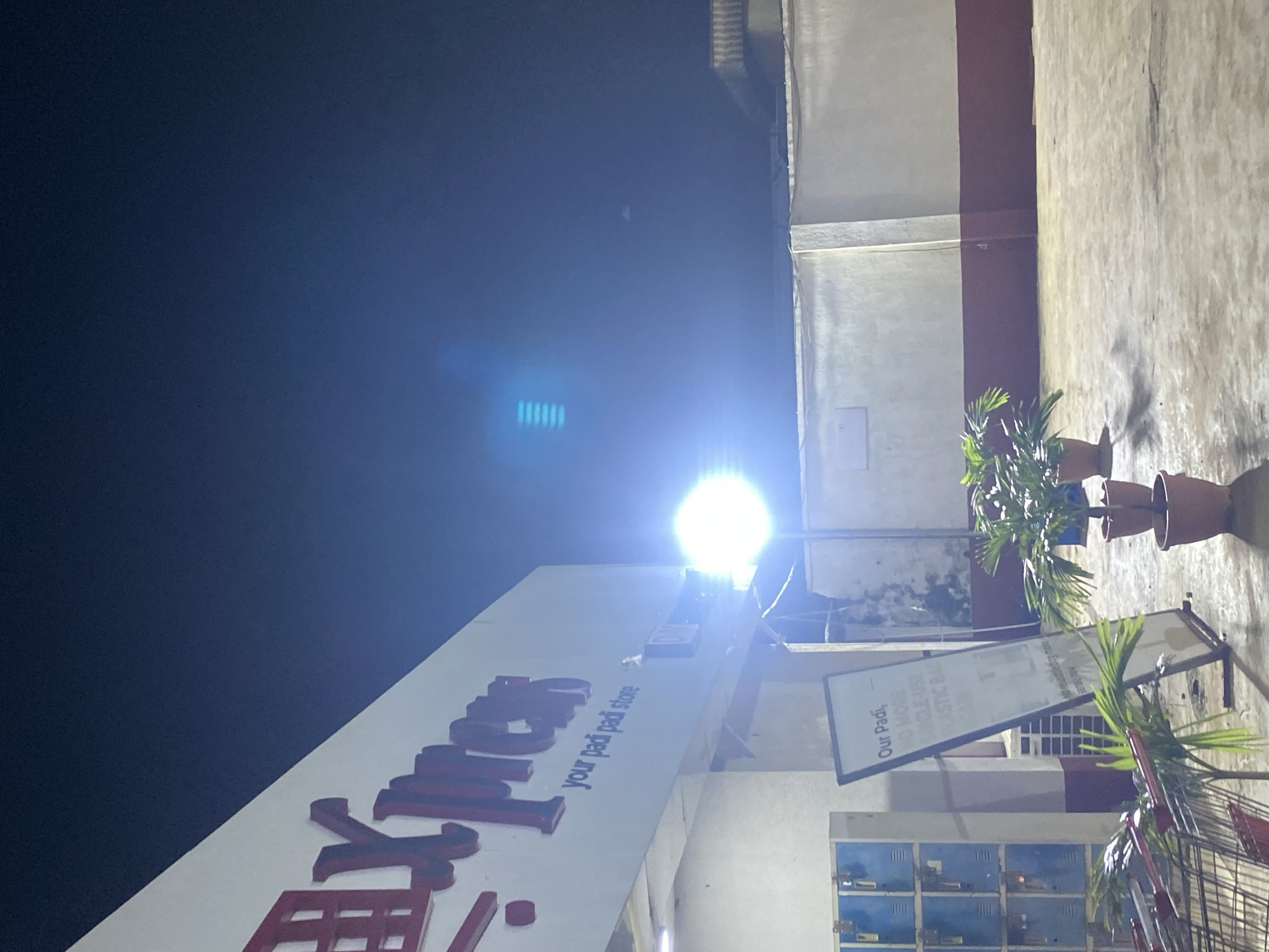 This nighttime image depicts the exterior of an "Xpress your padi padi store" located in Ikorodu, Nigeria. The building's white facade features a large red sign with a prominent stylized character, resembling "田", followed by "Xpress" and the tagline "your padi padi store" below. To the left of the main entrance, a stack of blue metal lockers is visible. An A-frame sign stands on the paved foreground, partially displaying the text "Our Padi, NO MORE SINGLE-USE PLASTIC BA". Shopping carts are present in the bottom-left corner of the frame.
The outdoor area is a light-colored paved surface, upon which several potted plants, appearing to be small palms, are strategically placed. A very bright, circular artificial light source is positioned in the mid-ground, generating a significant lens flare that includes a distinct blue rectangular artifact in the upper-left quadrant of the dark night sky. The background features a two-toned wall: a lower section painted dark red and an upper section in light beige. A partial view of a roofline with visible corrugations or ridges is present above the wall on the far right. No individuals are visible within the scene.