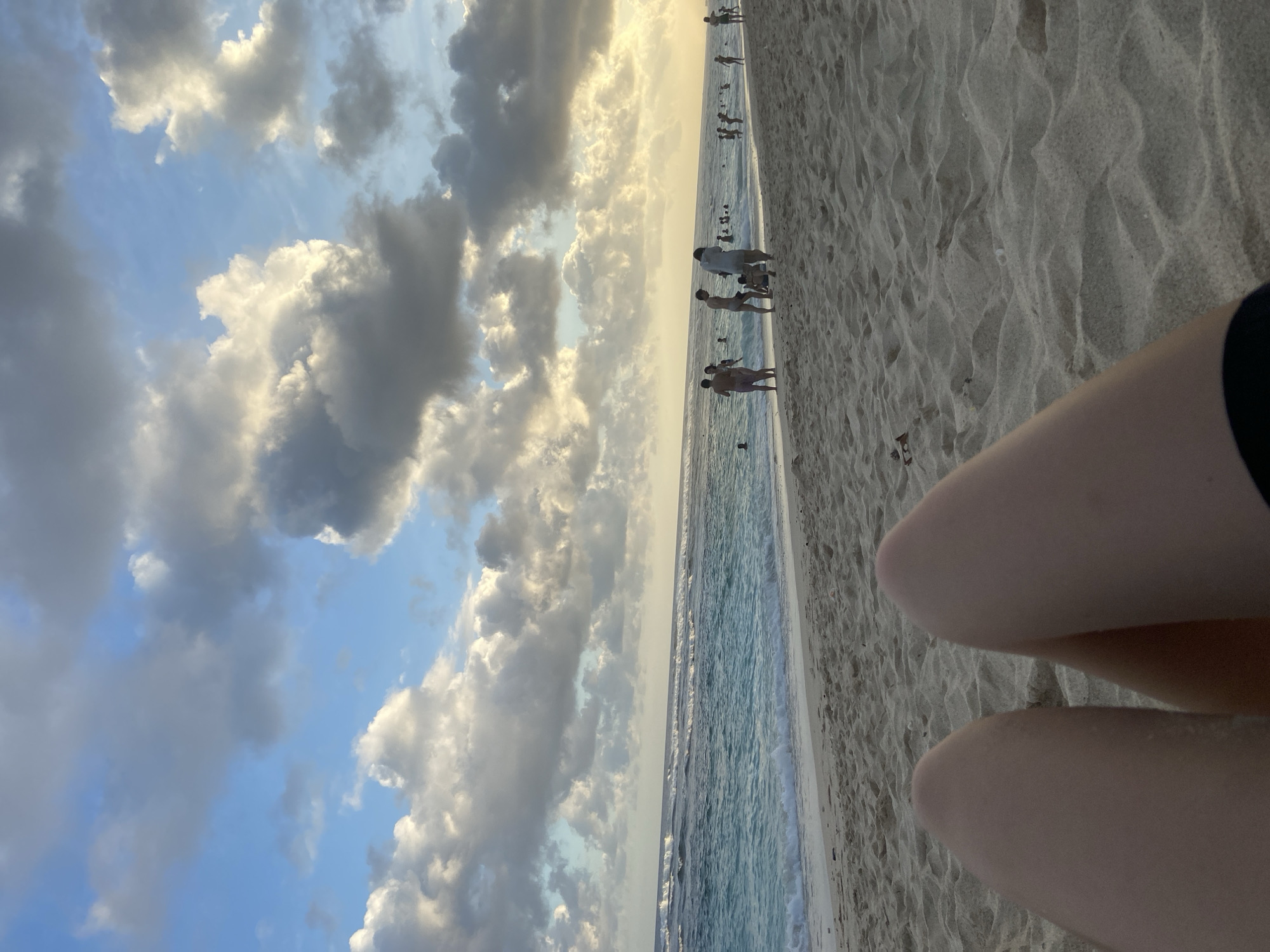 The image depicts a beach scene in Kuta Selatan, Indonesia. A portion of a person's legs is visible in the foreground, resting on the sand. The beach stretches into the distance with several people walking along the shoreline. The ocean meets the sand, and there are visible waves. Above, the sky is filled with numerous clouds, with patches of blue sky visible between them. The image is taken from a ground-level perspective.