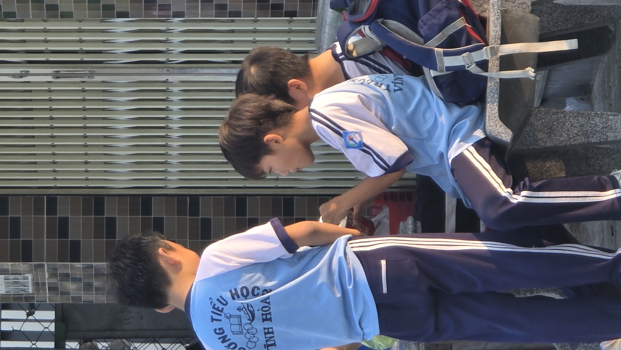 Three boys in school uniforms are interacting on a city street in Bắc Nha Trang, Vietnam. One boy is standing while the other two are crouched down next to him, possibly adjusting something. The boys are wearing light blue and white shirts and navy blue pants with white stripes. A school bag rests nearby on a concrete ledge. The background includes a tiled wall and a shuttered gate.