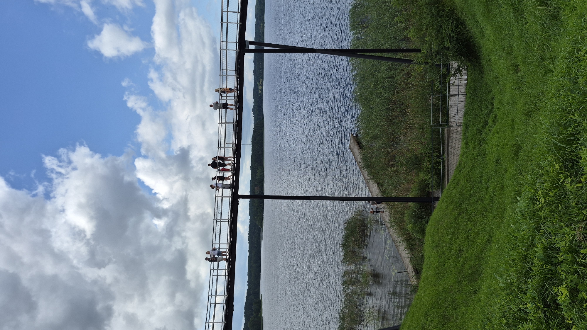 The image shows a pedestrian bridge over a lake in Zarasai, Lithuania. The bridge is made of metal and wood, and it is supported by two dark vertical poles. Several people are walking on the bridge, some of whom are looking at the lake or the surrounding scenery. The lake is calm and reflects the blue sky and white clouds. On the shore, there is a pier and some reeds. In the foreground, there is a grassy hill. The overall scene is peaceful and scenic, suggesting a pleasant day for outdoor activities.