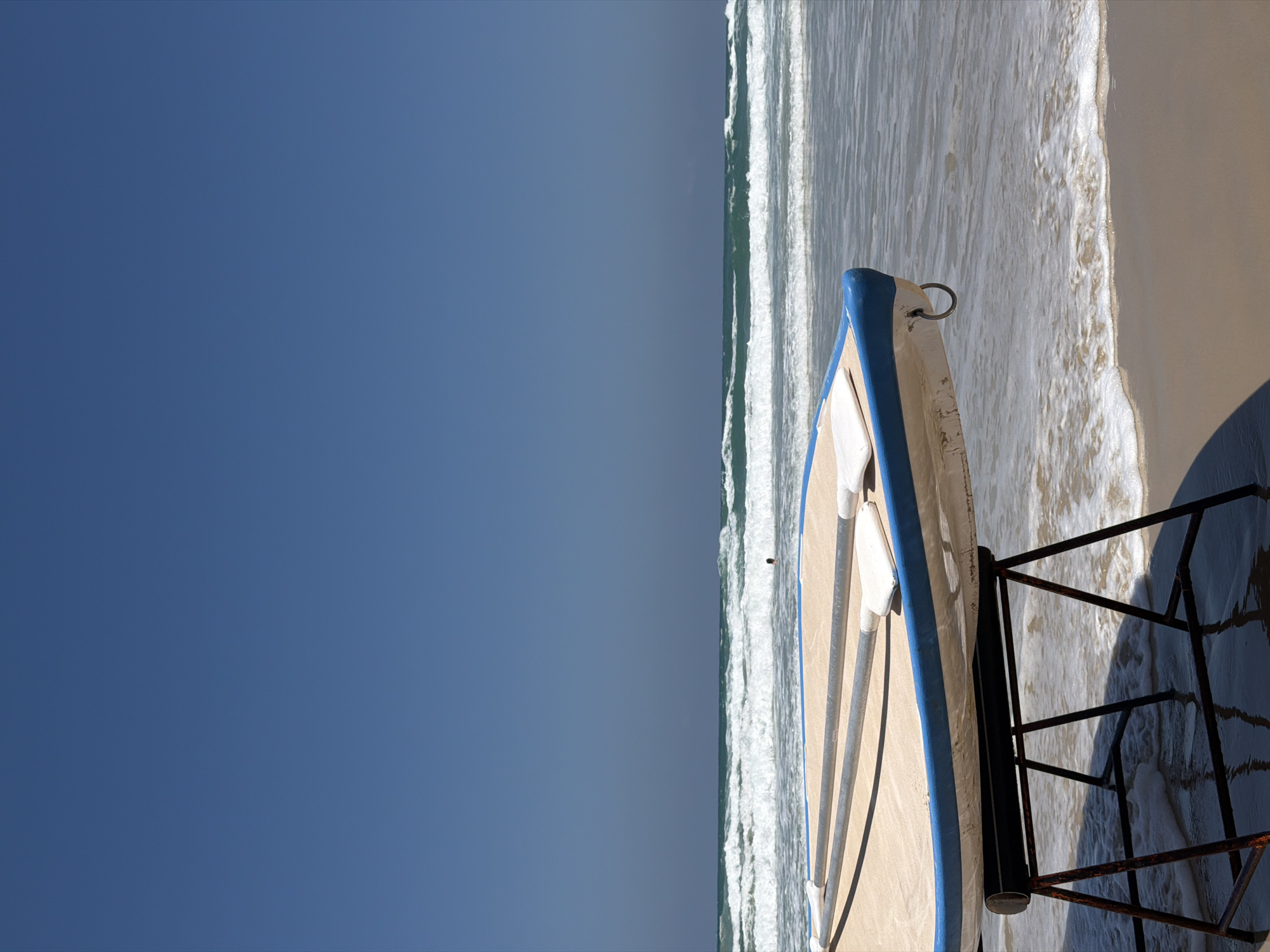 A high-angle shot depicts a partly visible beach scene in Bat Yam, Israel, focusing on a boat and its surrounding environment. The left side of the frame is dominated by a clear blue sky. The right side reveals the ocean with small, foamy waves breaking onto the sandy beach. A small boat, painted white with a blue trim, is stationed on the sand, partially submerged in the shallow water. A dark, rusted metal boat stand sits under the boat, partially submerged in the water. The image suggests a sunny day at the beach.