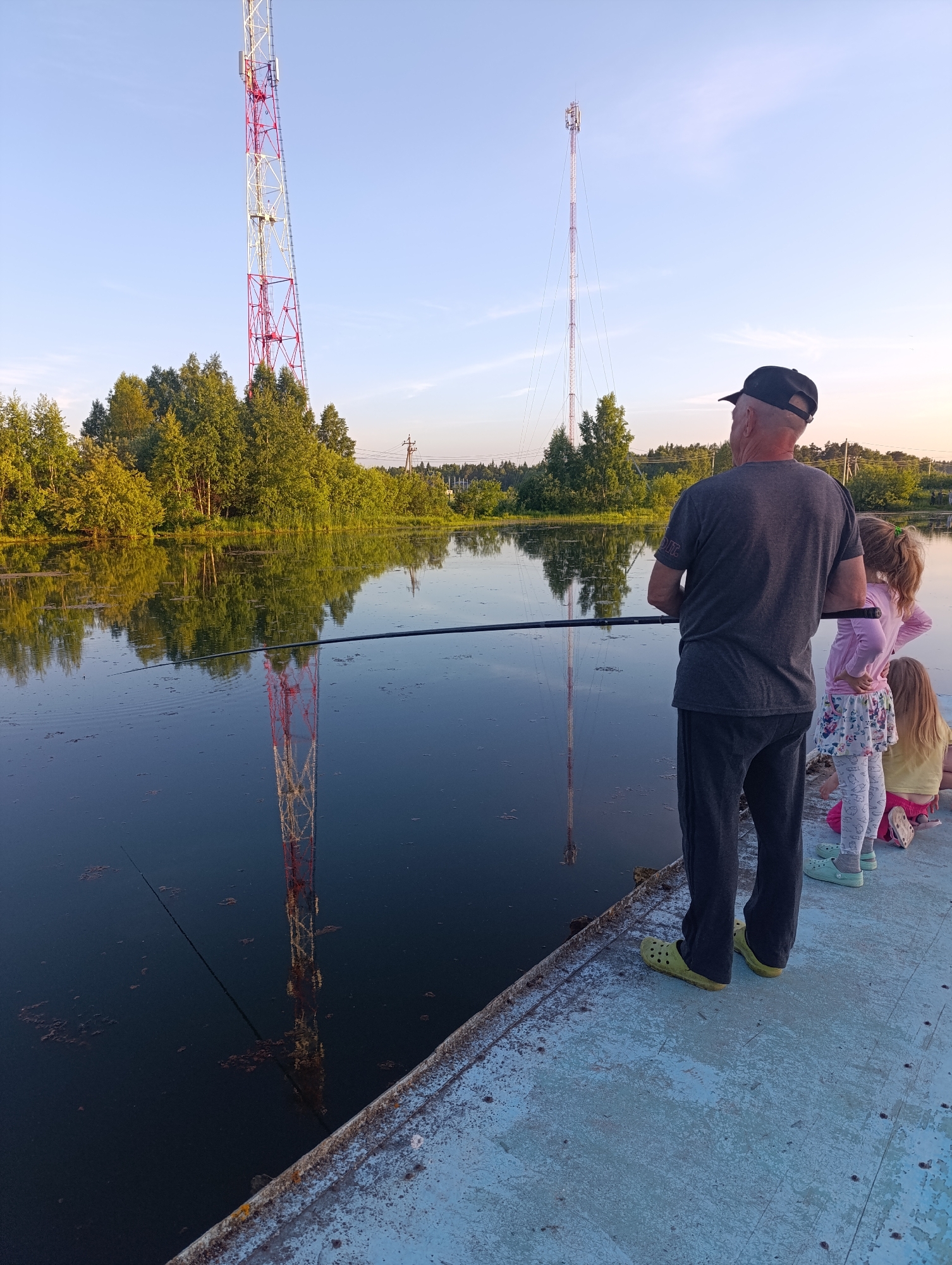 An outdoor scene depicts an adult male and two young children standing on a weathered concrete platform beside a calm body of water in an unknown city/location. The man, viewed from behind, wears a dark baseball cap, a grey t-shirt, dark trousers, and bright yellow-green Crocs, holding a fishing rod horizontally over the water. To his right, a taller child, likely a girl, wears a pink long-sleeved shirt, a white patterned skirt, and mint green Crocs, with her hands on her hips. A second, smaller child in a yellow t-shirt and pink shorts is crouched closer to the platform's edge.
The still, dark water prominently reflects the clear blue sky, which transitions to a light yellow hue near the horizon. Two distinct towers are reflected in the water and stand against the sky in the background. The tower on the left is a red and white lattice structure with visible antenna arrays. The tower on the right is a taller, slender guy-wired mast, also equipped with communication antennas at its apex. Dense green trees line the far bank of the water, and power lines with utility poles are visible above the tree line in the distance on the right. The concrete platform is light blue-grey, showing signs of chipped paint and wear.