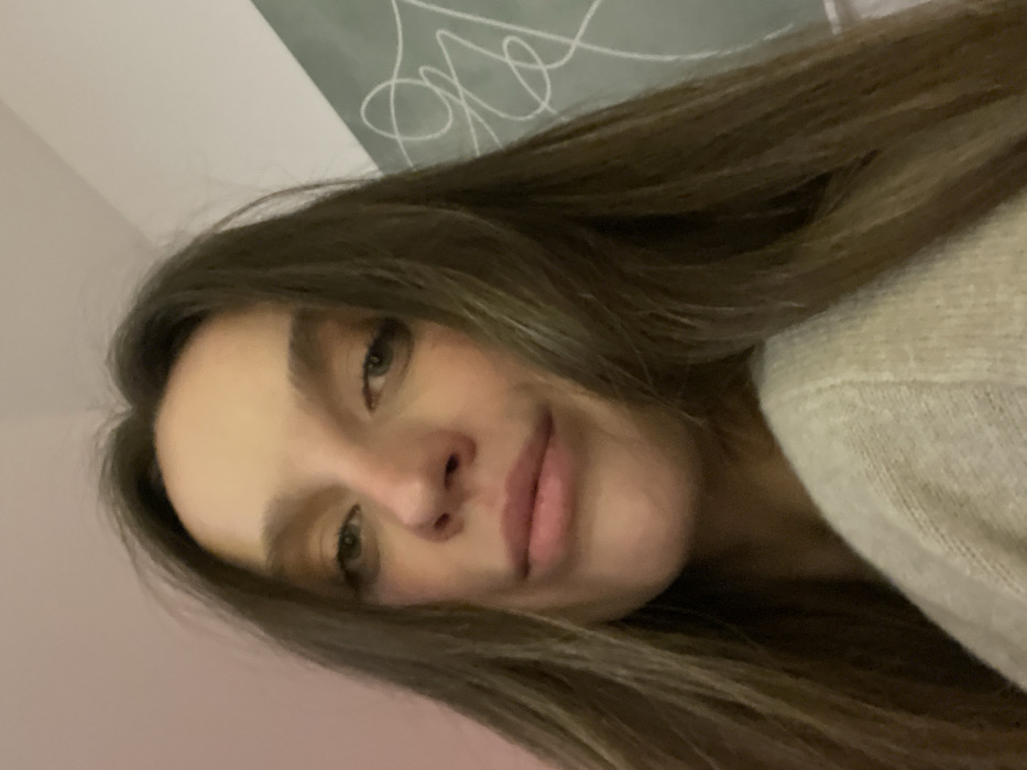 A young woman's face is captured at a tilted angle. She has light olive-toned skin, brown hair, and green eyes. She is wearing a cream-colored sweater. Her expression is soft and she appears to be slightly smiling. In the background, there is a light-colored wall with what appears to be light gray scribbles or abstract designs. The overall lighting is soft and diffused.