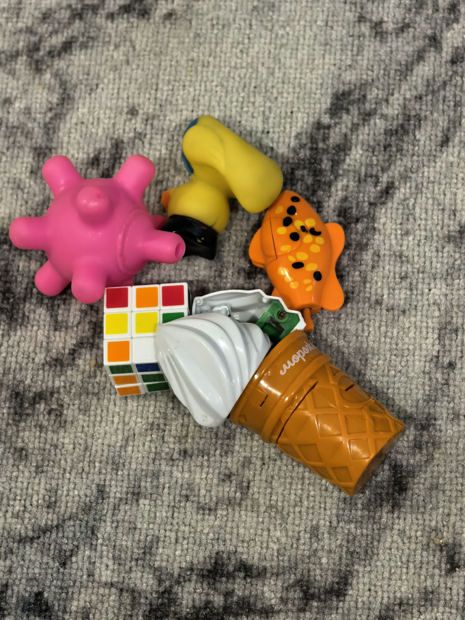The image shows an assortment of children's toys on a gray carpet with dark gray swirls. The toys include a pink, possibly rubber, spiky ball; a yellow and black plastic toy; an orange toy fish with black spots; a partially solved Rubik's Cube; and a plastic toy ice cream cone, orange with white "ice cream." The ice cream cone appears to be broken or disassembled. The carpet is evenly lit. There is no indication of the city of Sterlitamak, Russia, in the image.