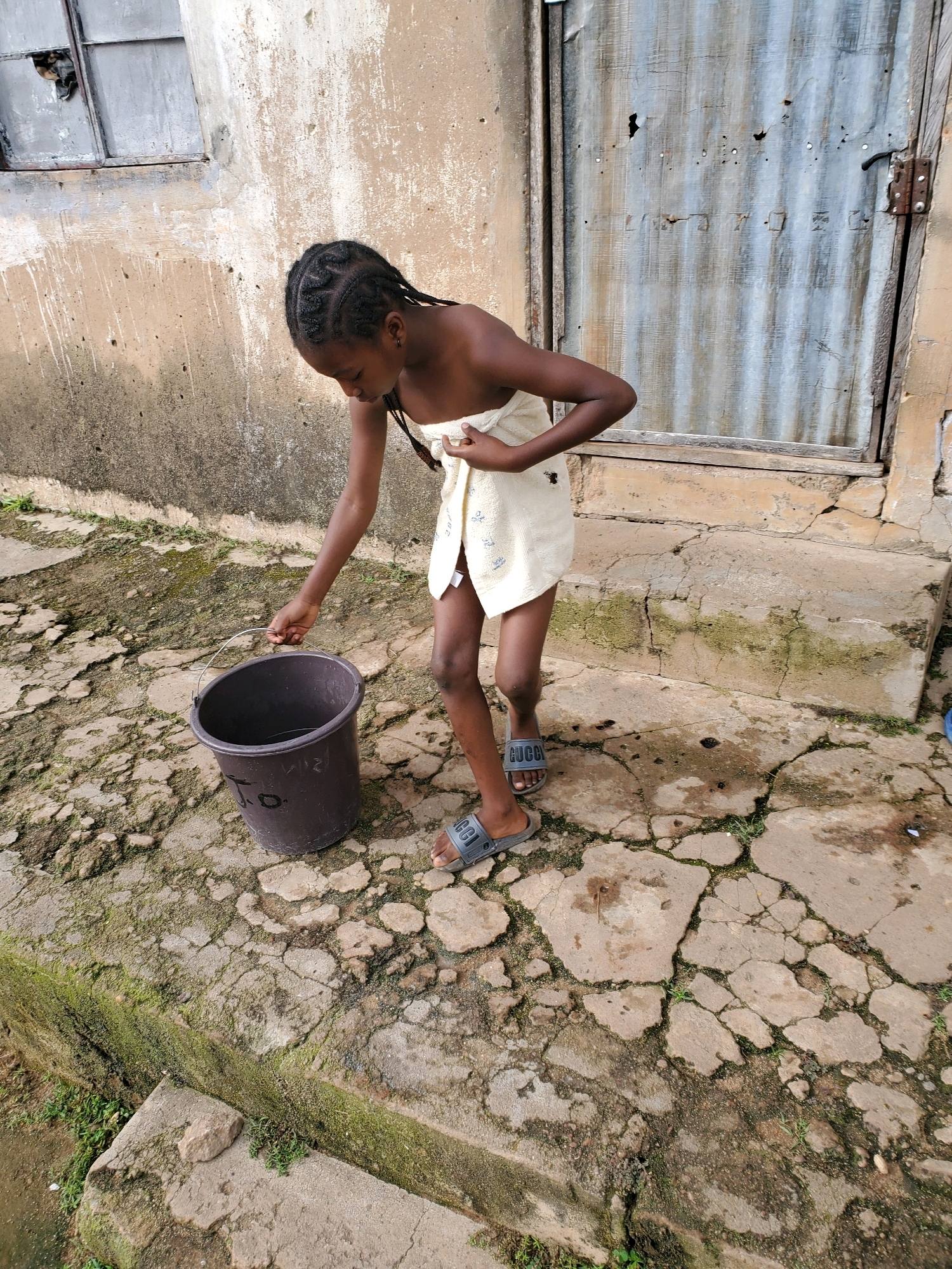 A young girl with braided hair, wrapped in a white towel, stands in an outdoor courtyard in Jos, Nigeria. She is holding the handle of a dark bucket with her right hand, dipping it into a shallow pool of water on the ground. The ground is paved with cracked concrete and patches of moss and weeds. Behind her, a weathered building with a corrugated metal door and a damaged window is visible. The scene appears to depict a moment of daily life, possibly related to water collection or bathing.