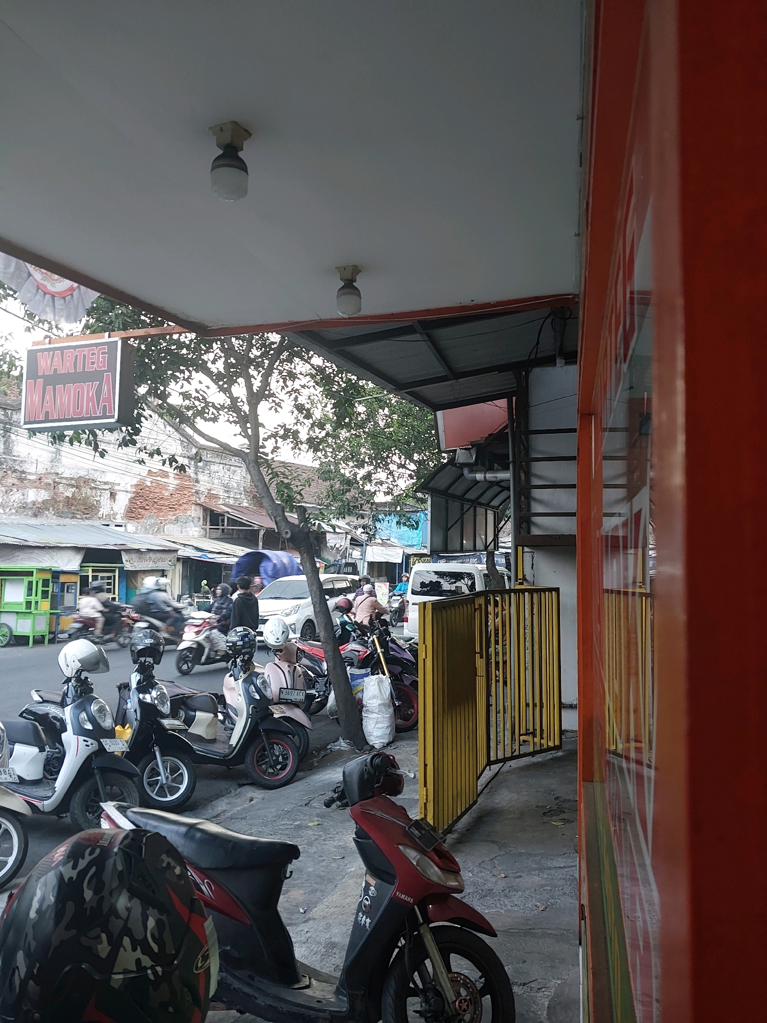 The image depicts a street scene, likely in the city of Malang, Indonesia, with a focus on a row of parked motorcycles and a building with an orange facade. The foreground shows several scooters parked along a sidewalk. One scooter in the lower left is partially obscured by a camouflage-patterned helmet. Further down the sidewalk, other scooters are visible, some with riders or helmets on them.
Behind the motorcycles, a street is visible with a few cars and motorcycles in motion, suggesting moderate traffic. A sign with "WARTEG MAMOKA" in red lettering is prominently displayed overhead. The buildings lining the street appear to be a mix of old and newer structures. To the right, a bright orange wall with vertical white lines dominates the frame, likely part of a shop or commercial establishment. A yellow metal gate is also visible, providing access to an area behind it. Trees with dense foliage are interspersed throughout the scene, casting shadows. The lighting suggests it is daytime, possibly late afternoon or early morning, given the soft light.