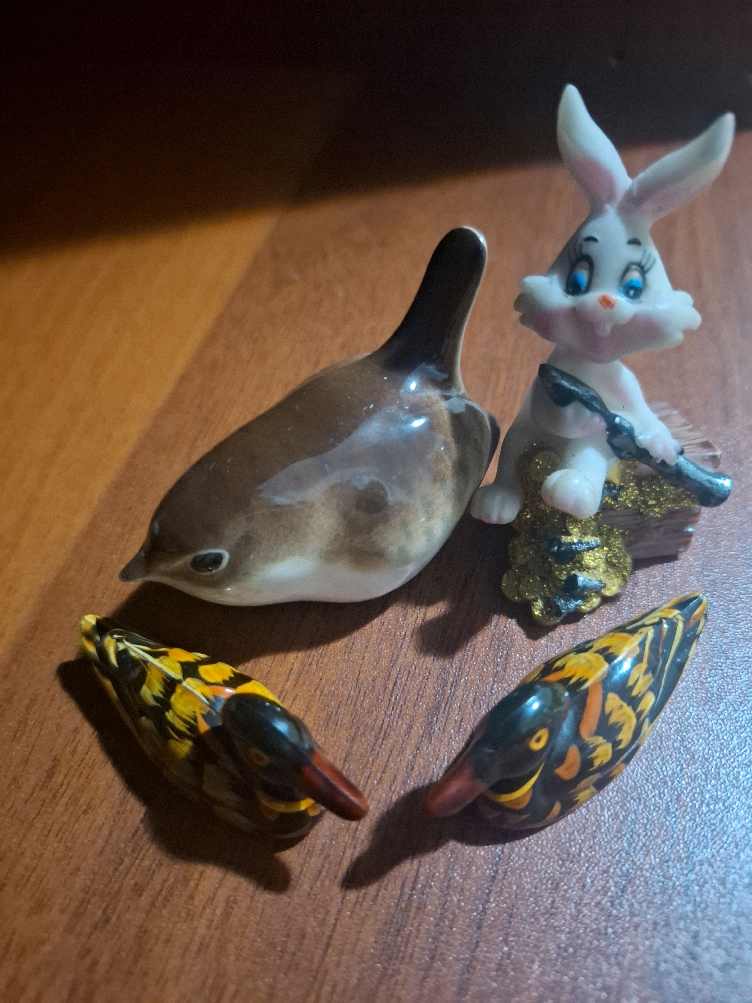 Four ceramic figurines are arranged on a wooden surface. In the foreground are two small, brightly painted duck figurines, one with yellow, black, and orange patterns, and the other with similar colors but a more prominent orange beak. Behind these ducks, a larger, brown and white ceramic bird figurine is positioned on its side. To the right of the bird is a white ceramic rabbit figurine with blue eyes and pink accents, holding a black object that appears to be a brush or stick. The rabbit is adorned with gold glitter at its base. The wooden surface appears to be a tabletop with a warm, brown tone and visible wood grain. The lighting in the image suggests an indoor environment, with shadows cast by the objects. The scene depicts decorative figurines. There is no information in the image that allows for the identification of a specific location in the city of Almaty, Kazakhstan.