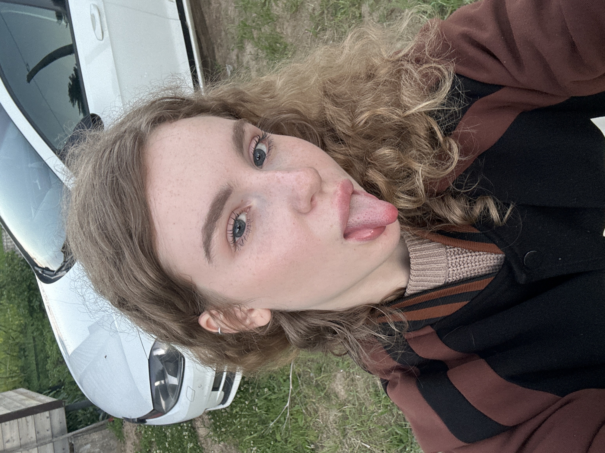 A young individual with long, curly, light brown hair and blue-grey eyes is captured in a close-up selfie, rotated 90 degrees counter-clockwise. They have visible freckles across their nose and cheeks and are playfully sticking their tongue out towards the camera. A small silver hoop earring is visible in their left ear. They are wearing a black jacket with prominent brown stripes on the sleeves and collar, layered over a beige knitted sweater.
In the background, a white car is partially visible, showing its front bumper and side, parked on what appears to be gravel or dirt. Green grass and sparse vegetation are also seen on the ground. The scene suggests an informal outdoor setting, potentially a yard or driveway in Vasilyevo, Russia. The lighting is soft and even, indicating daytime, possibly late afternoon or overcast conditions. The individual's expression conveys a playful and lighthearted mood. There is no visible text in the image.