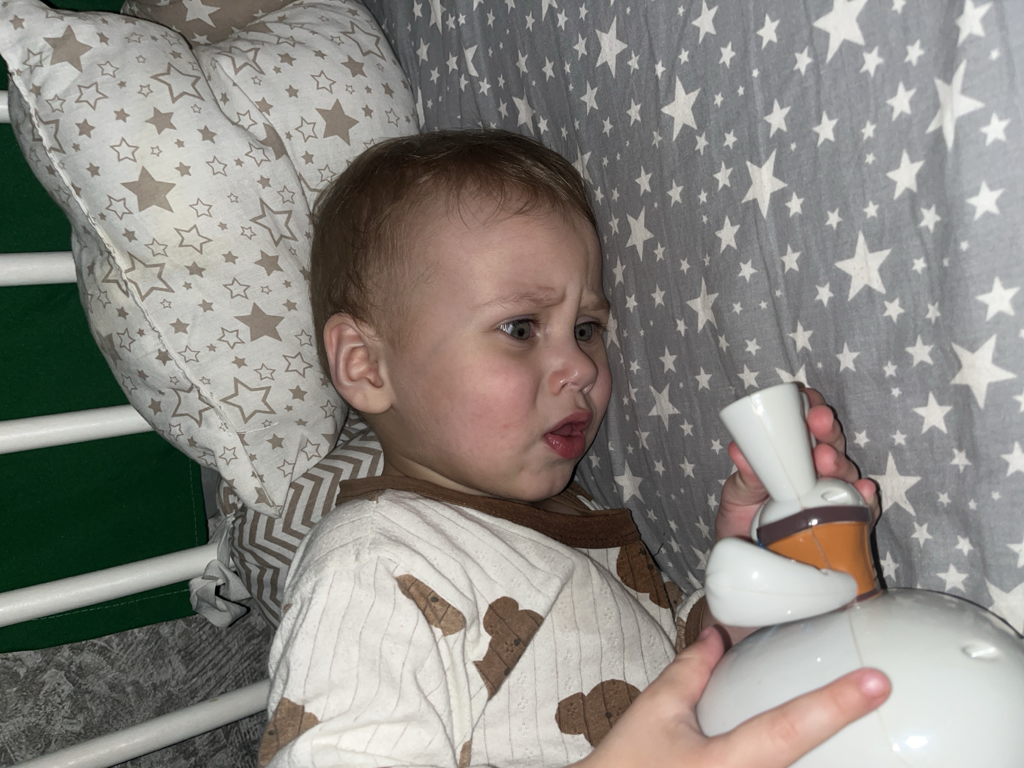 A young child lies in a crib or bed, holding a white and orange toy, possibly a lamp. The child has a worried or surprised expression. A patterned pillow is behind the child, and a gray blanket with white stars is in the background. The crib has white rails and a green side. The location in the city of Фоки, Russia is not evident from the image.