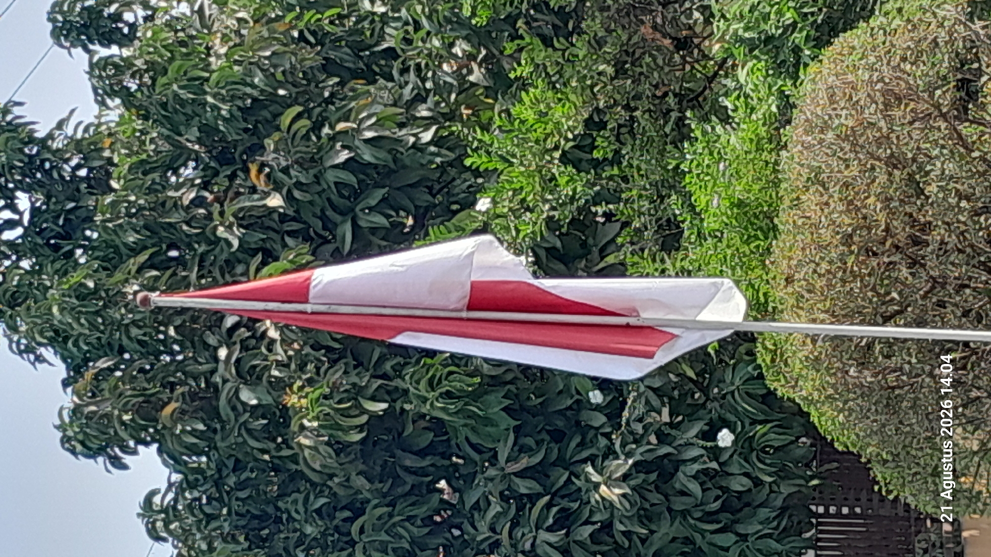 The image shows an Indonesian flag, red and white, flying from a flagpole. The flag is partially unfurled and appears to be slightly creased. The flagpole is positioned in front of a dense backdrop of green foliage, likely trees and bushes. In the lower left corner, a timestamp is visible: "21 Agustus 2026 14.04". This suggests the photograph was taken in Takeran, Indonesia, on August 21, 2026, at 2:04 PM. The environment appears to be outdoors, possibly a garden or park, given the presence of trees and bushes. No people are visible in the image.