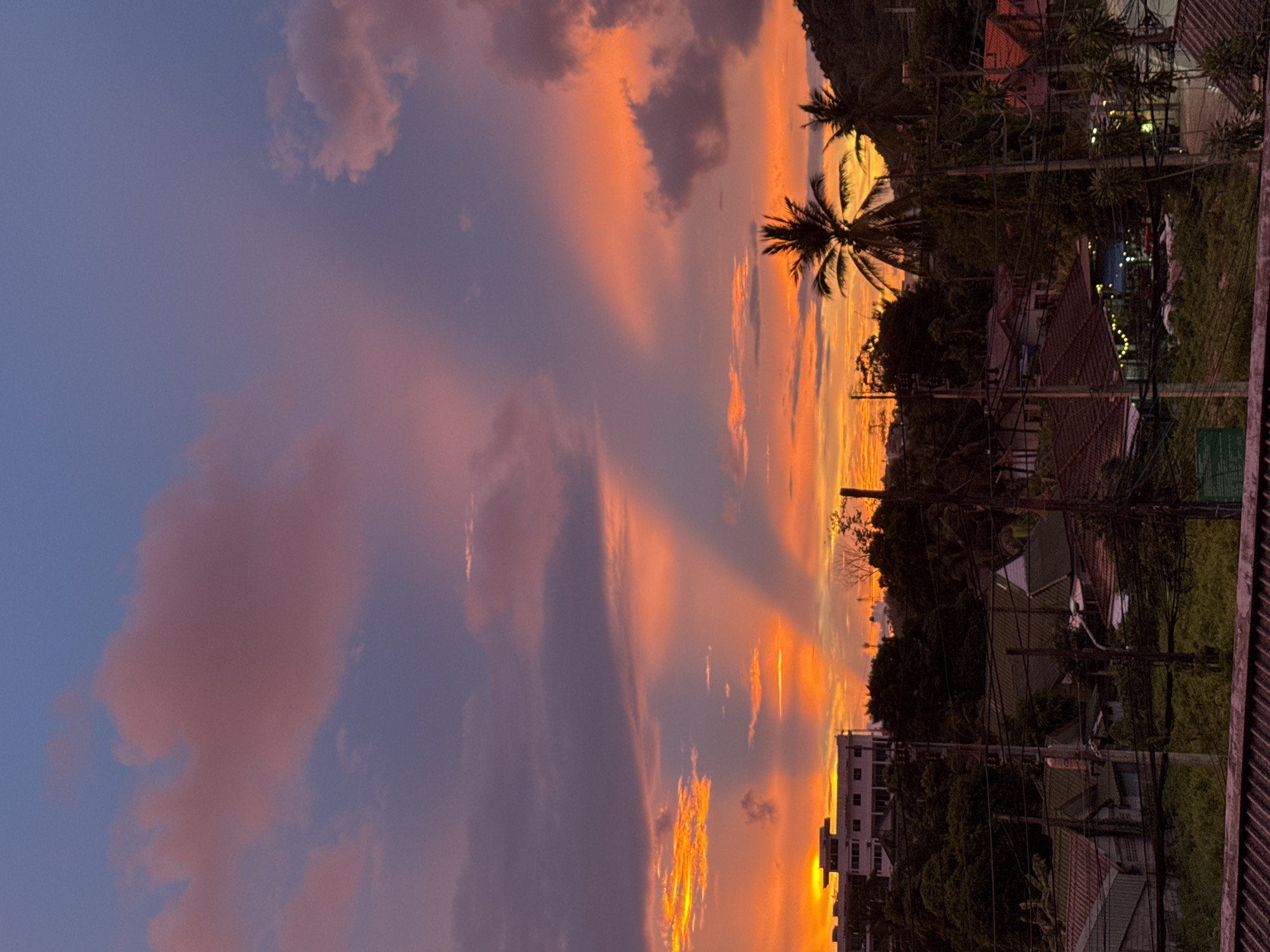 The image depicts a vibrant sunset over the city of Karon, Thailand. The sky is a dramatic canvas of orange, pink, and purple hues, with crepuscular rays emanating from behind scattered clouds. In the foreground, a low-rise urban landscape is visible, characterized by residential buildings with terracotta tiled roofs, lush green trees, and a complex network of electrical wires and poles. A multi-story white building stands prominently on a hillside, overlooking the other structures. Two palm trees are silhouetted against the colorful sky, suggesting a tropical setting. The overall scene is tranquil, capturing the beauty of nature as day transitions into night in a populated area.