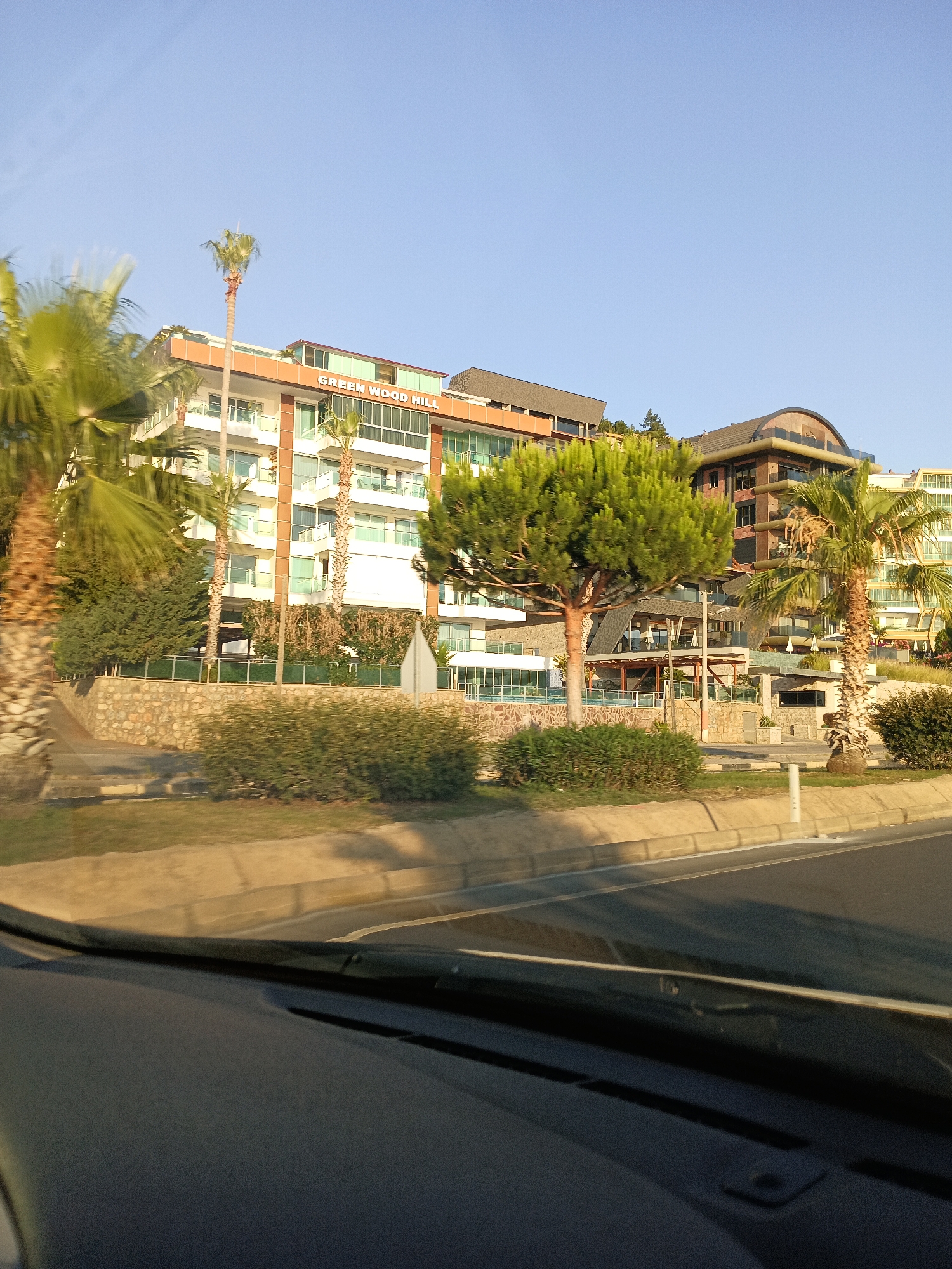 The image shows a view from a car, looking out through the windshield at a modern hotel complex. The hotel, identified by the sign "GREEN WOOD HILL", features multiple stories with balconies and large windows. It is surrounded by lush greenery, including palm trees and manicured bushes. In the foreground, there is a road with a sidewalk and a low stone wall. The sky is clear and blue, indicating a sunny day. The architecture suggests a resort or residential building located in Alanya, Turkey.