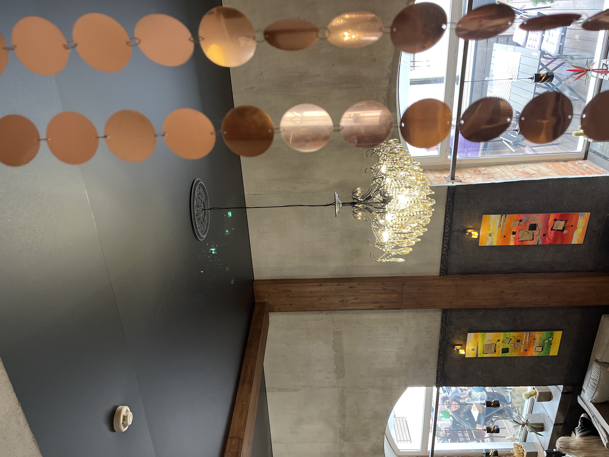 The image shows an interior scene, likely a cafe or restaurant, looking upwards towards the ceiling and windows. A dark ceiling is visible with a smoke detector on the left and a decorative ceiling rose above a chandelier. Two decorative strands of circular, copper-colored discs hang from the ceiling. A large, crystal chandelier is lit and suspended in the center of the frame.
The walls are a mix of rough concrete and dark, textured material. Two tall, vertical wooden beams are prominent structural elements. On the dark textured wall, two vertical abstract paintings with bright colors are displayed.
Large windows with arched tops look out onto an outdoor area with tables and chairs, suggesting the presence of a patio or balcony. Through the windows, people can be seen sitting at tables.
In the foreground, a glimpse of furniture and a person with long blonde hair sitting on a couch is visible. Plants in concrete pots are placed on a windowsill. The overall impression is a stylish, contemporary interior with a bohemian or eclectic decor. The location is identified as Vitsebsk, Belarus.