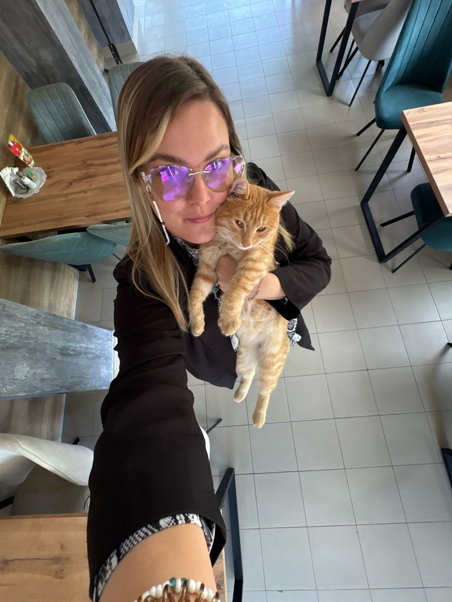A blonde woman wearing glasses and a black blazer is holding an orange tabby cat in her arms. The cat is looking to the left of the frame. The woman is holding the cat with her right arm and supporting its body with her left arm. The scene is indoors, with tiled floors and wooden tables and chairs visible in the background. The lighting suggests an indoor setting with ambient light. The overall impression is a casual indoor moment. The location is stated to be Noviy, Russia.